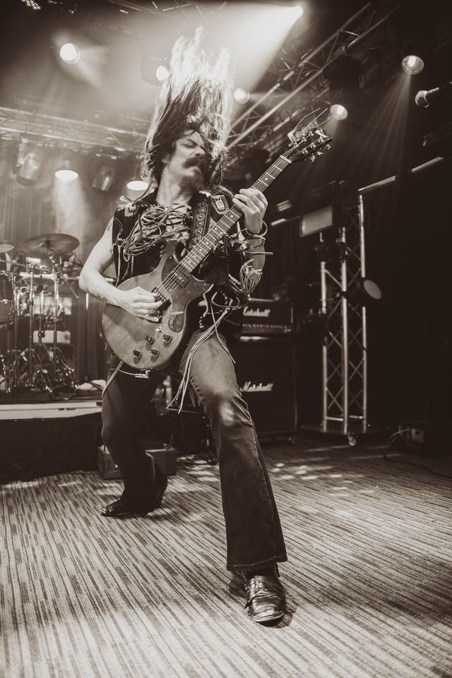 Guitarist performing passionately on stage, hair flying, in black and white concert photo with stage lights and musical equipment in background.