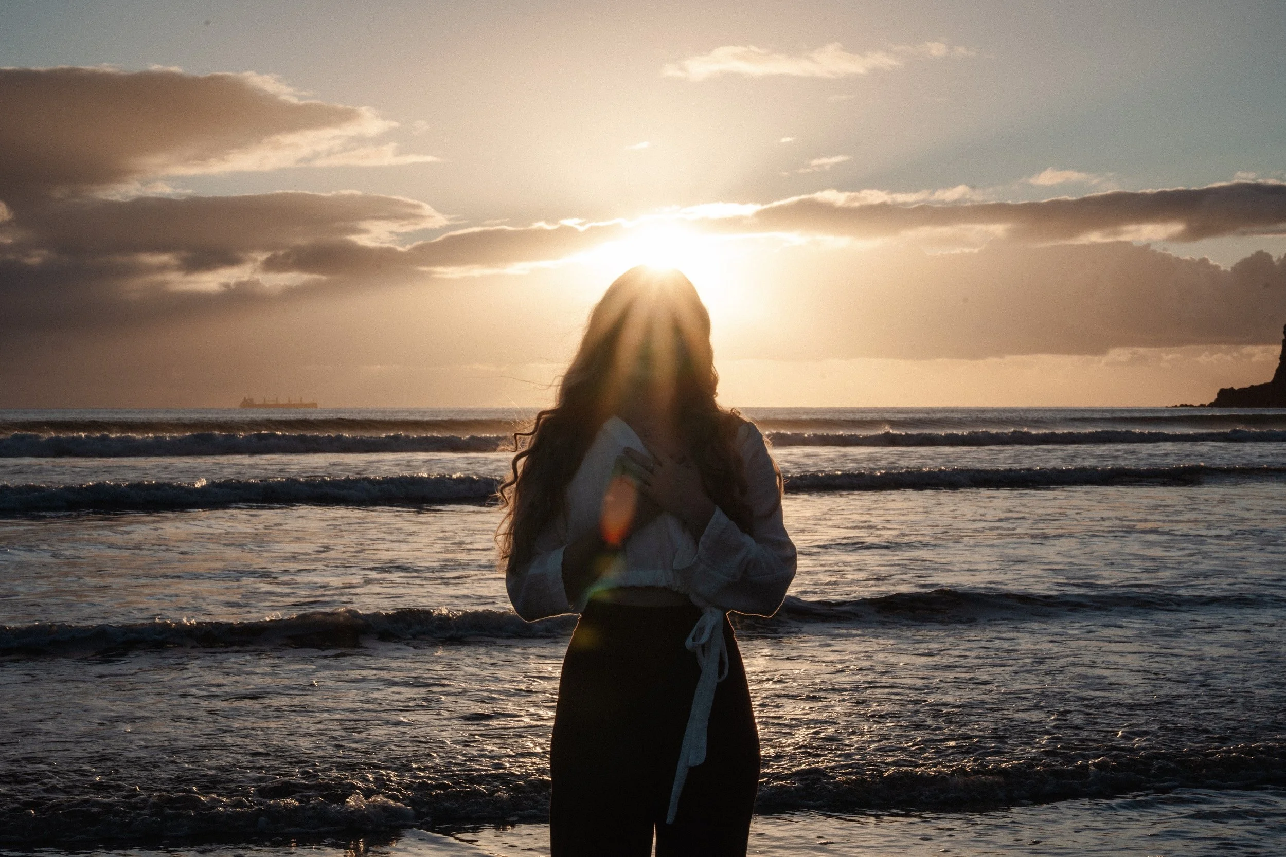 Silhouette photo of Sandrina from New Narratives standing on the beach in front of the sunrise, facing the camera and with her hands on her heart