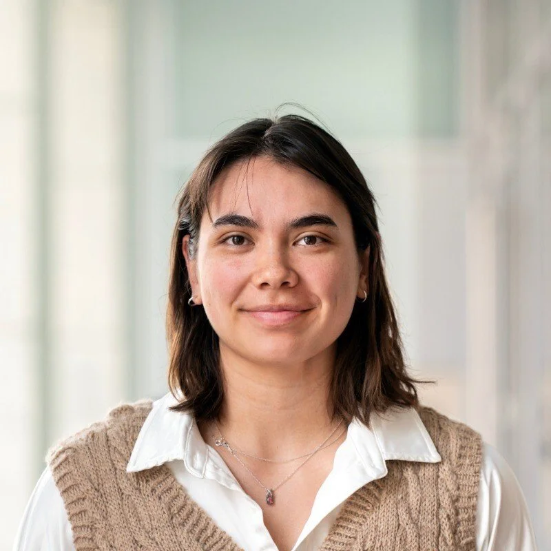 A woman smiling and wearing a white shirt, a cable-knit sleeveless sweater, small earrings, and a necklace, standing indoors with blurred background.