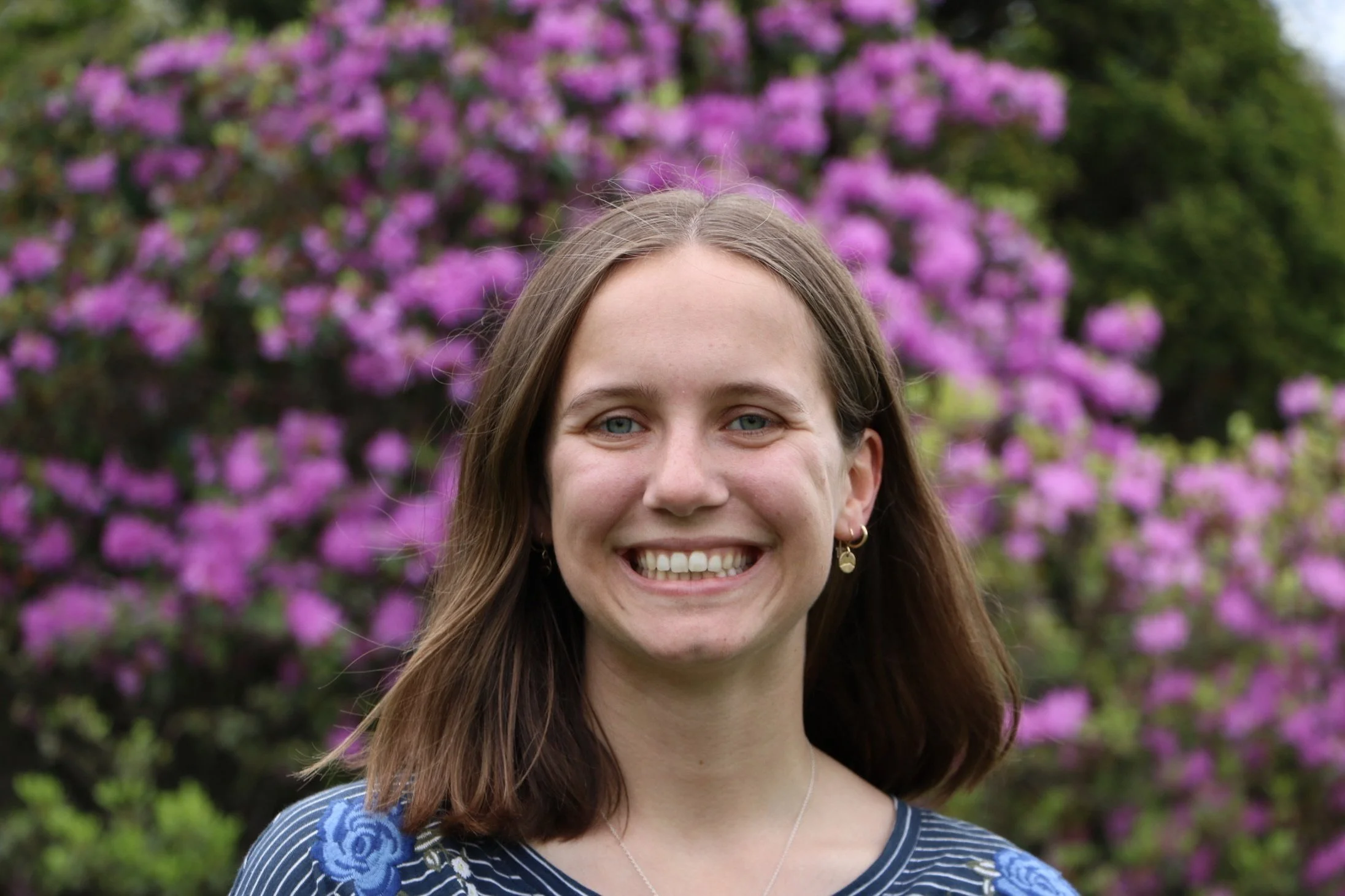 A woman smiling at the camera, standing outdoors in front of a blooming pink and purple flowering tree.