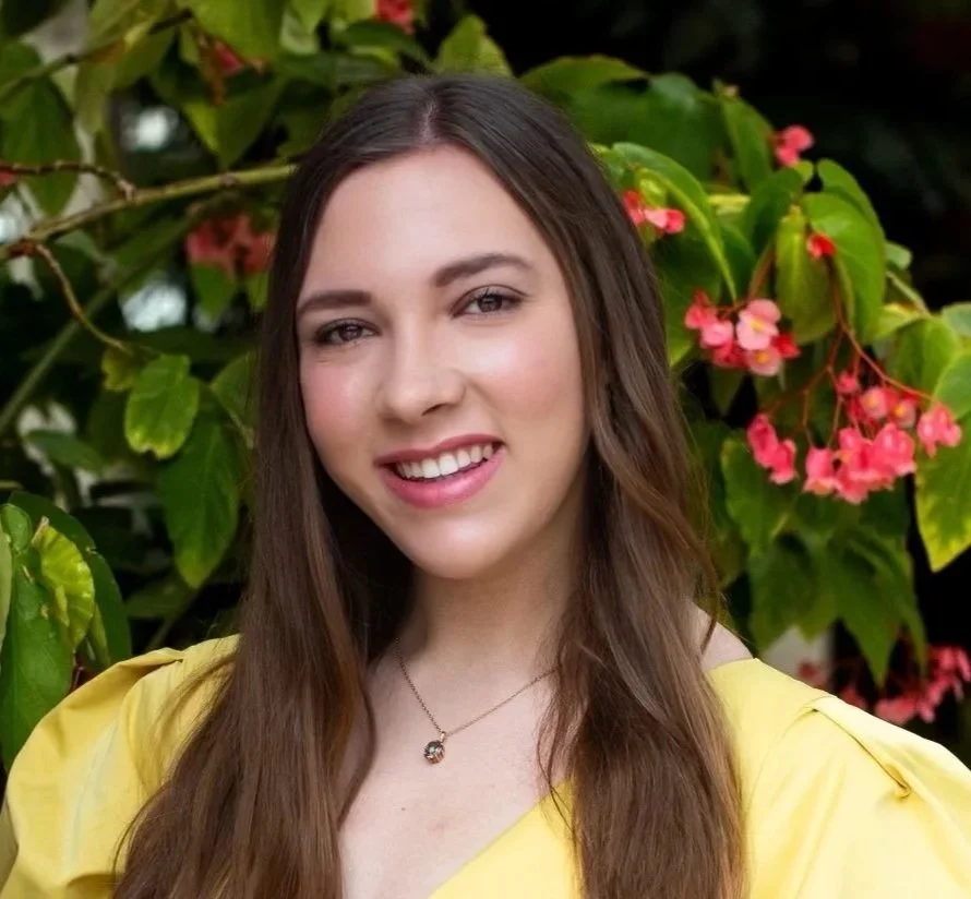 A smiling woman wearing a yellow top and a necklace, standing outdoors in front of green leaves and pink flowers.