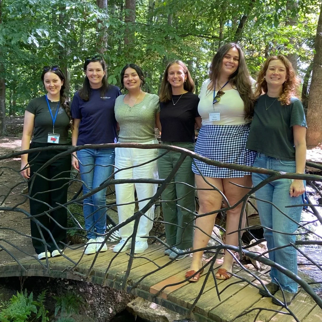 Group of seven women standing on a small wooden bridge in a lush green forest, smiling and posing for the photo.