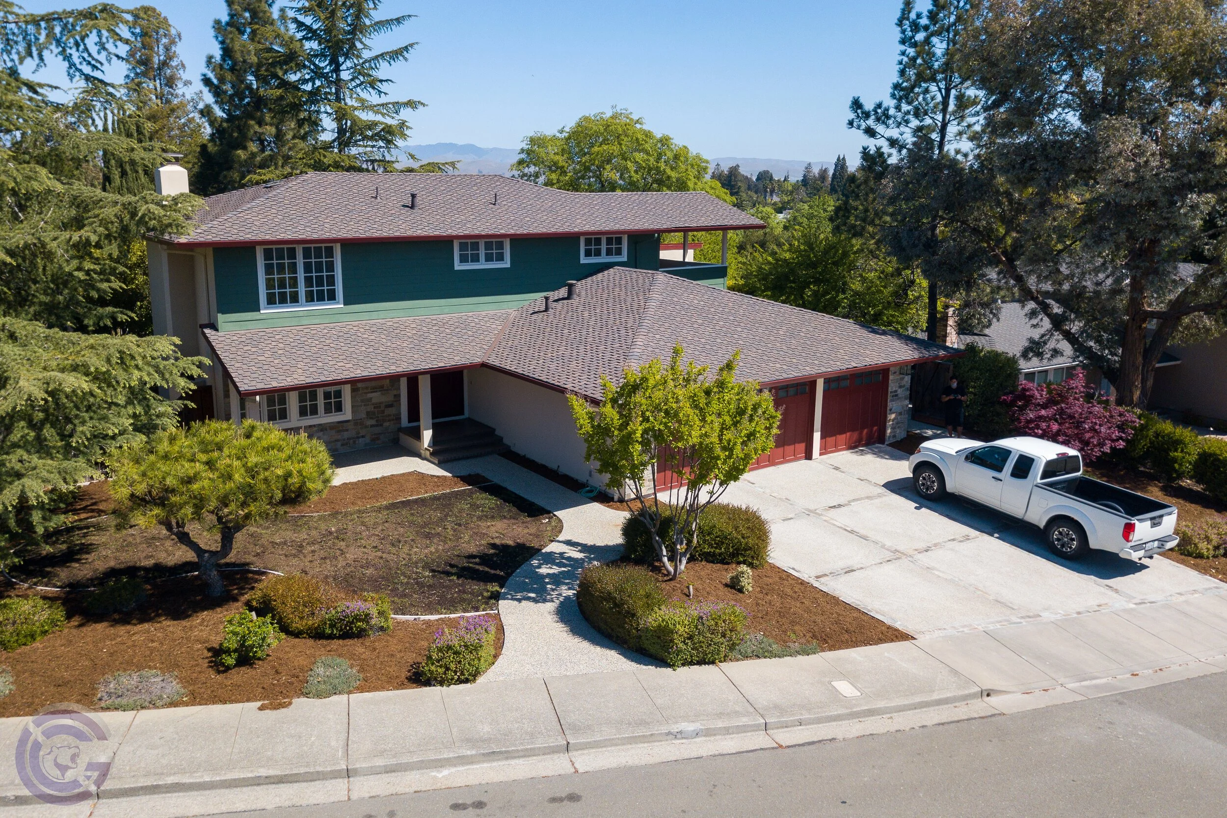 A two-story house with a green upper level, gray roof, and a red garage door, surrounded by trees and a landscaped yard, with a white pickup truck parked in the driveway.
