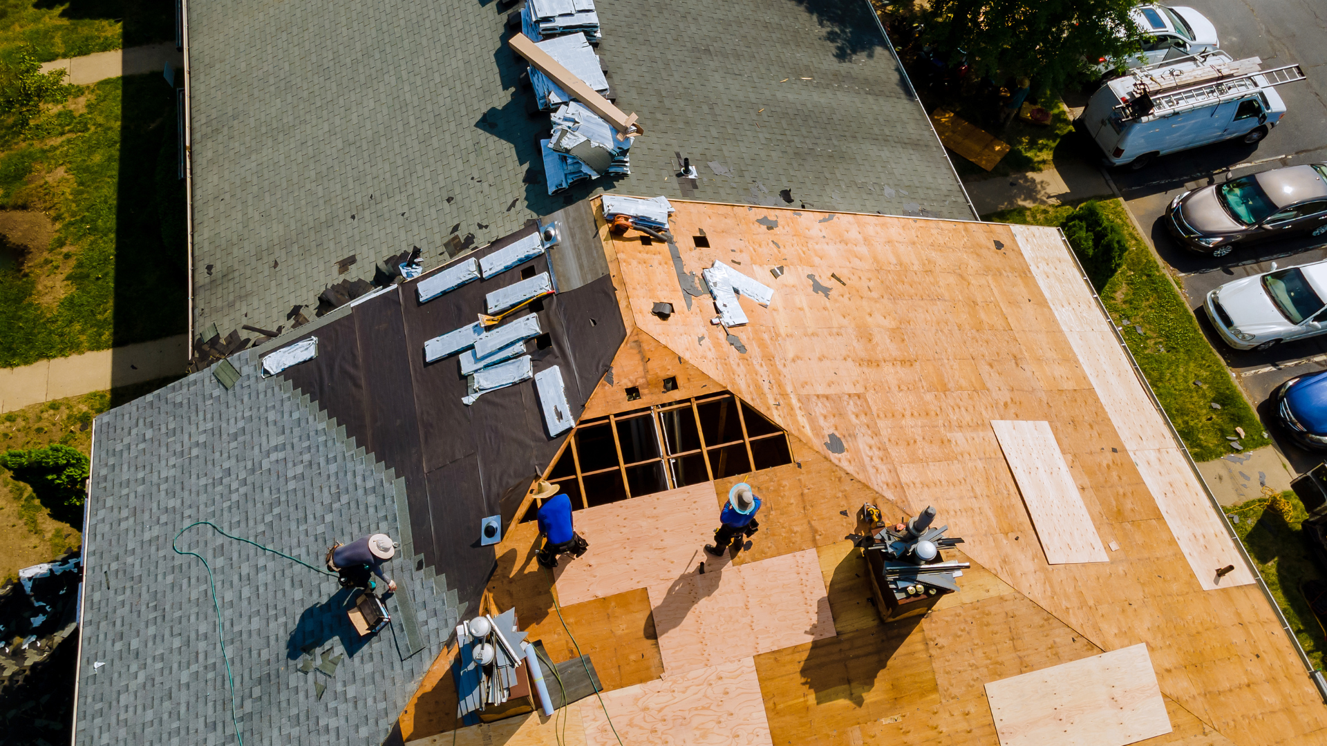 Aerial view of a house under renovation showing workers installing new roof panels. Some sections have existing shingles and others are covered with new plywood sheathing. The work area is surrounded by parked cars and a grassy area with trees.