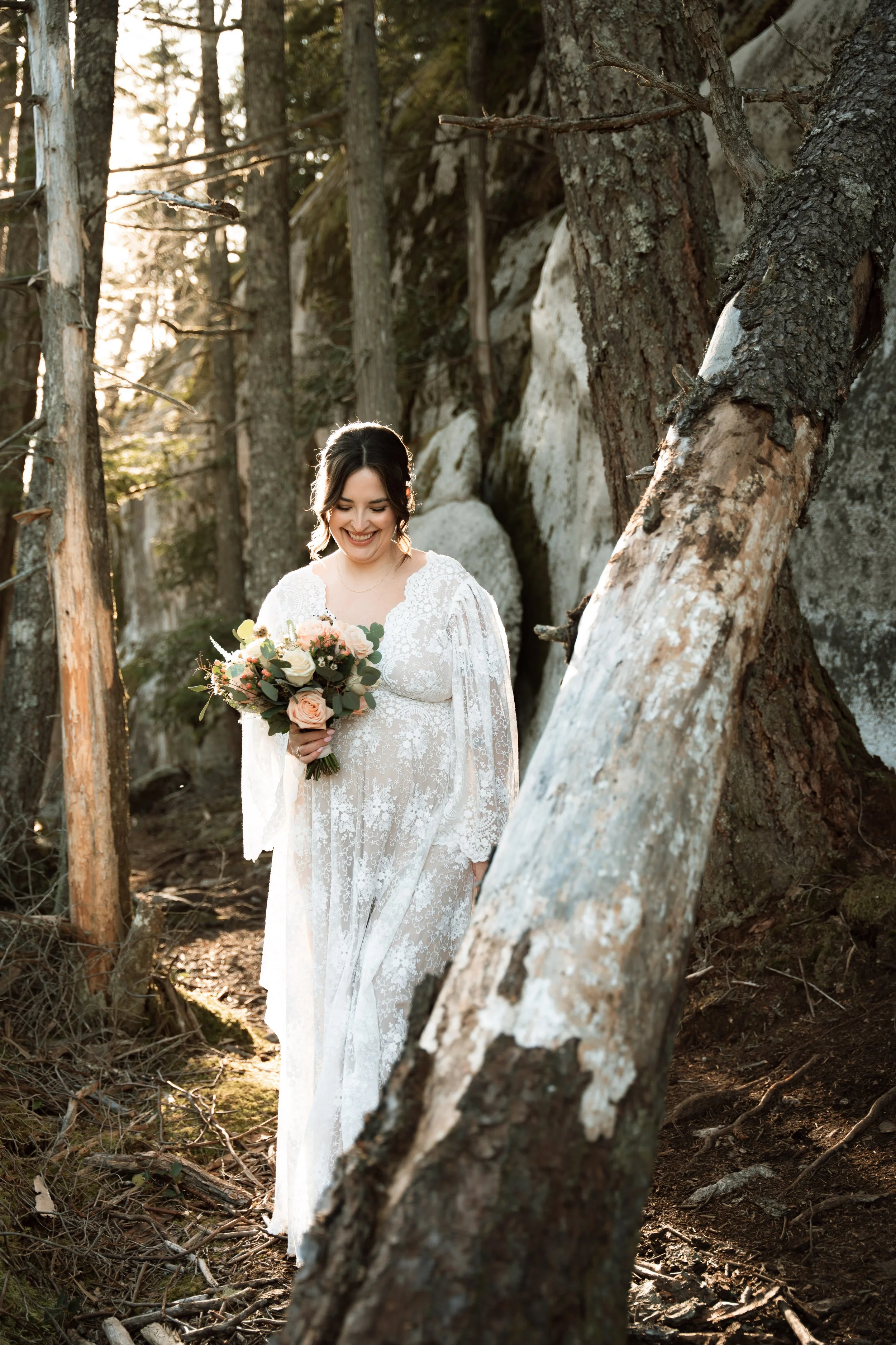 A woman in a white lace wedding dress holding a bouquet of pink and white roses, smiling in a wooded outdoor setting with tall trees and rocks, during sunset.