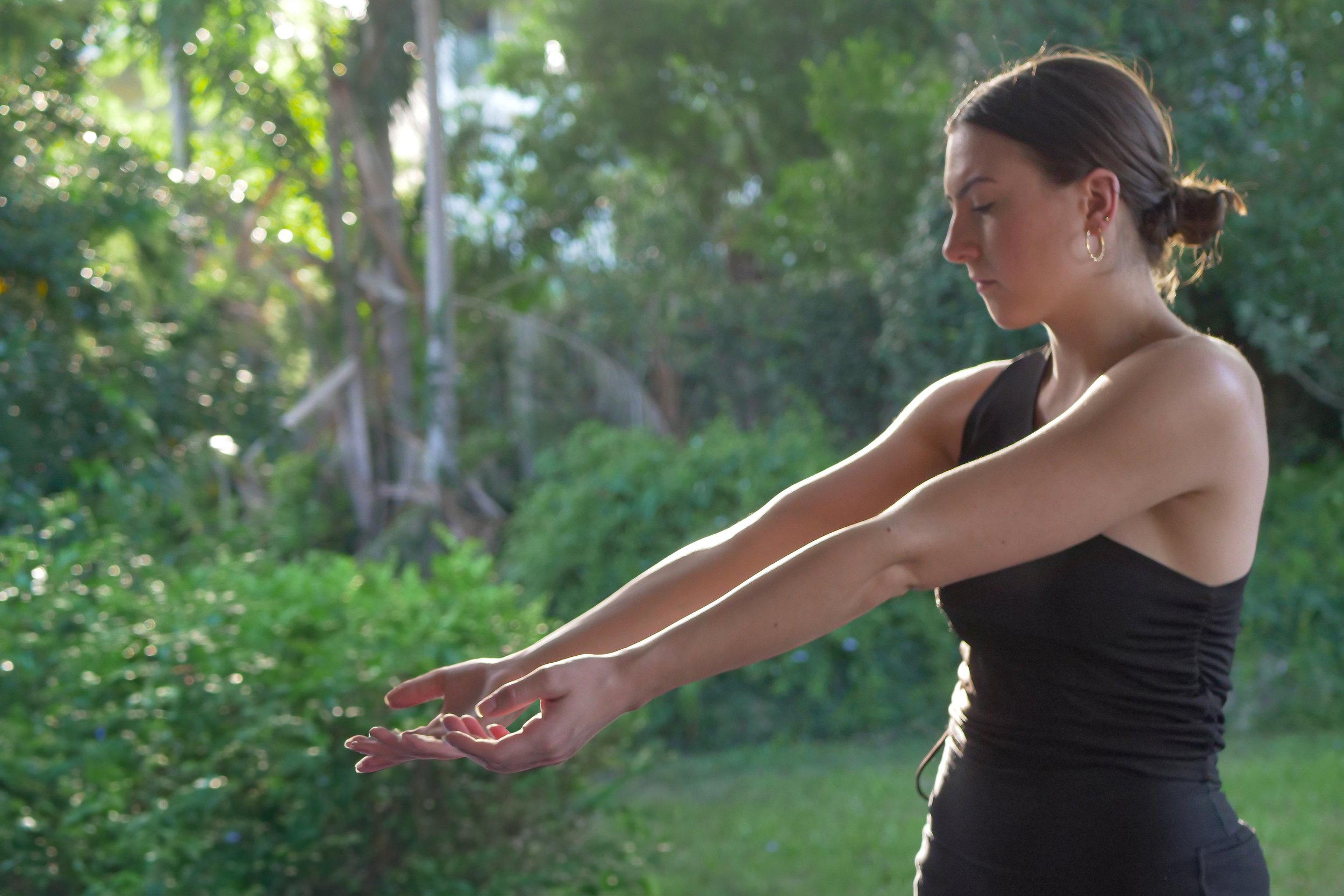 A woman in athletic attire extending her arms forward outdoors with green trees and sunlight in the background.