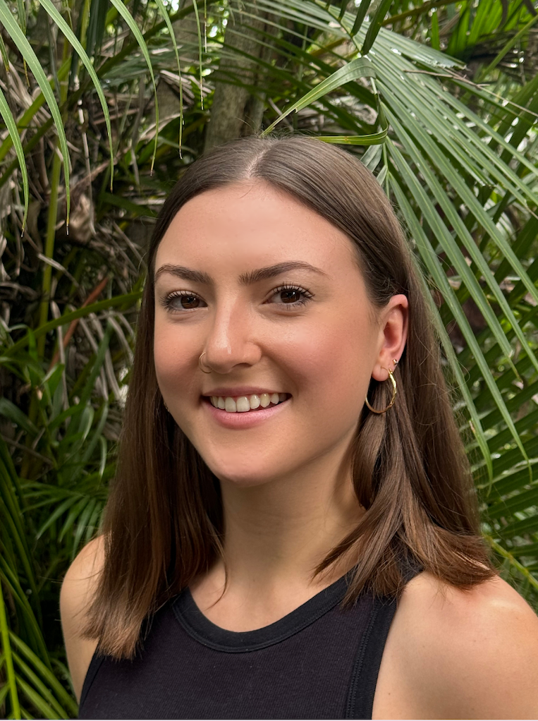 A young woman with shoulder-length brown hair, light skin, wearing hoop earrings and a nose ring, smiling at the camera outdoors among green tropical plants.