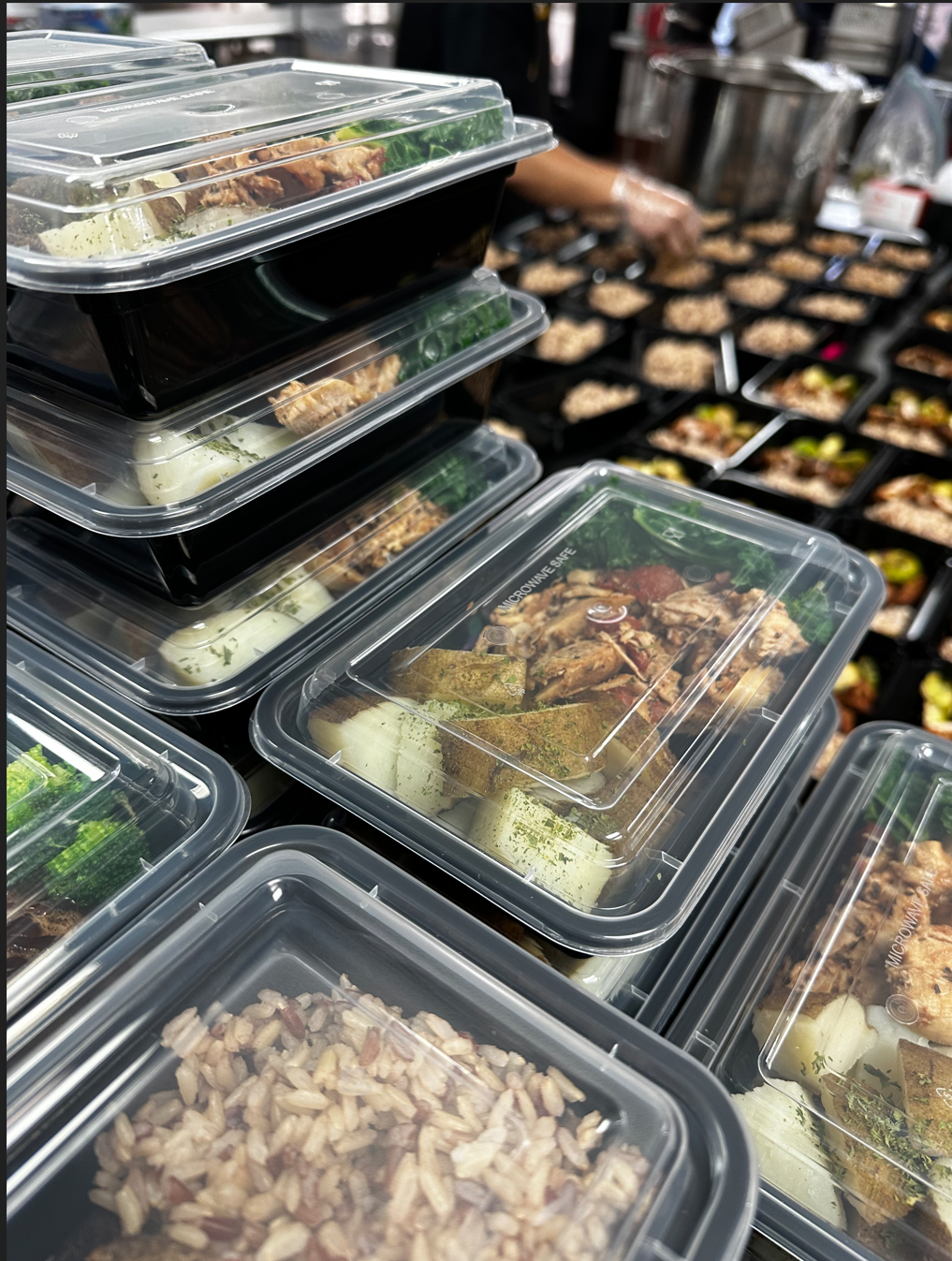 Stacked meal prep containers with cooked rice, vegetables, and meat in a grocery or food market setting.