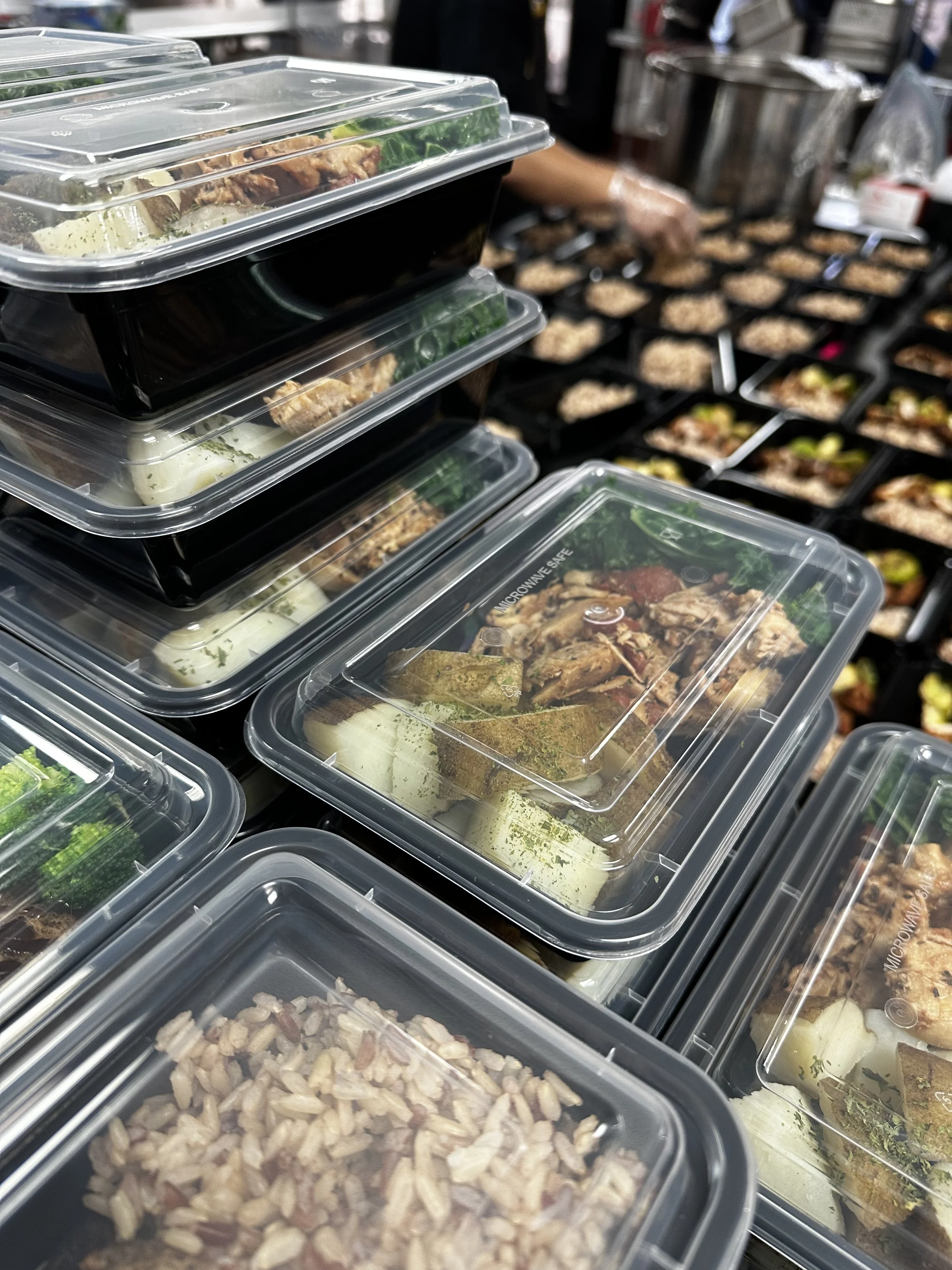 Clear plastic containers filled with prepared food items, stacked on shelves, with a busy kitchen or food prep area in the background.