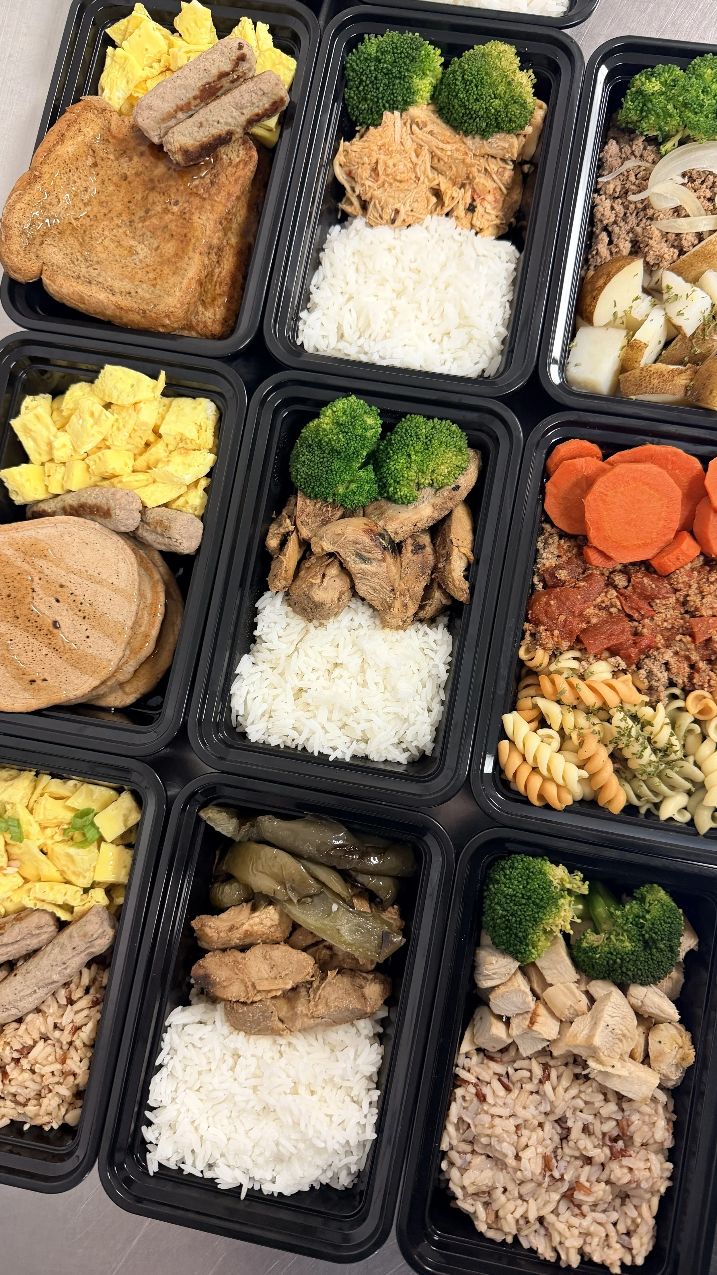 Multiple black meal prep containers filled with various foods including scrambled eggs, breakfast sausage, pancakes, broccoli, shredded chicken, carrots, pasta, ground beef, rice, and grilled chicken.
