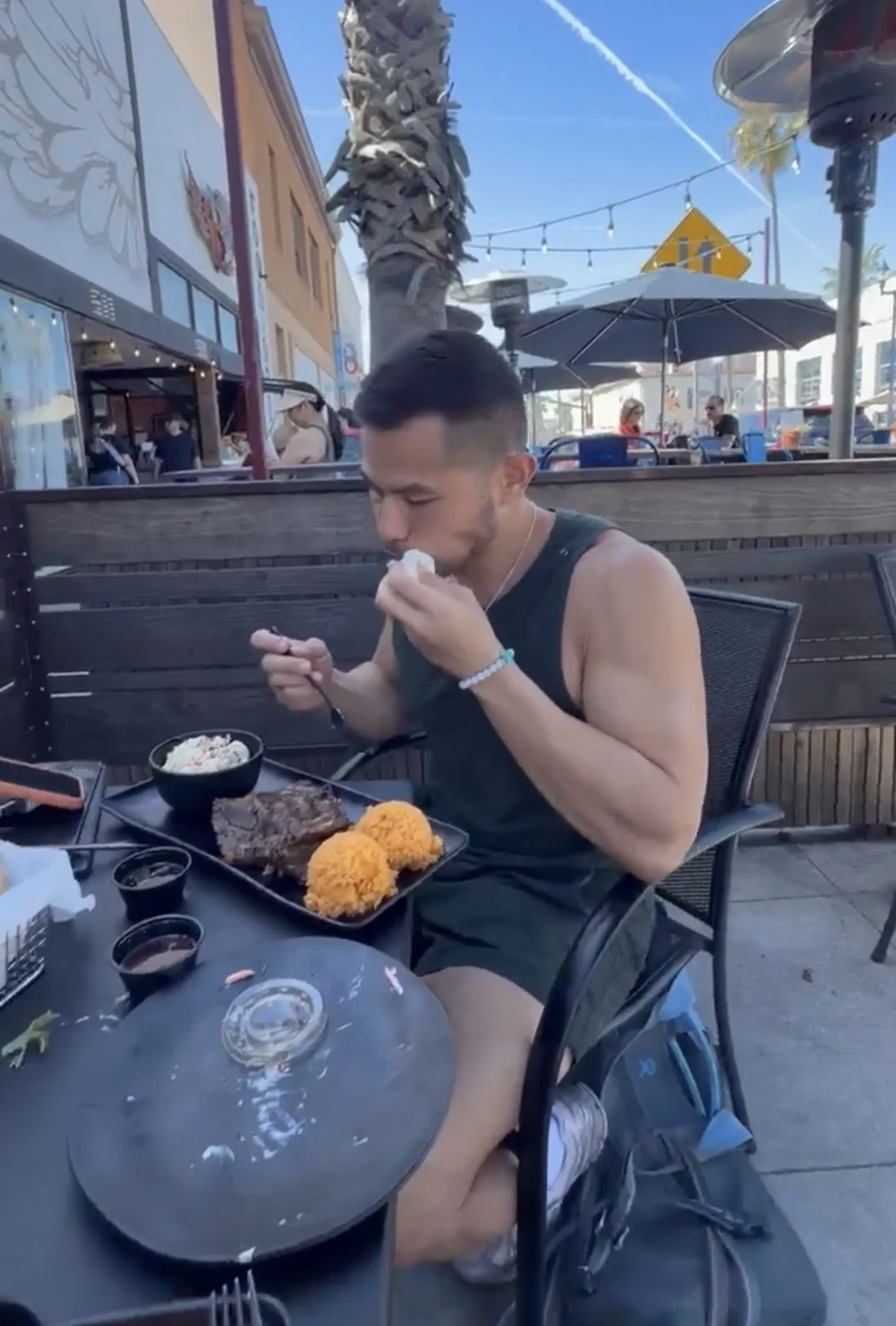A man sitting outdoors at a restaurant table, eating with a spoon, with plates of fried food, rice, and sauce in front of him. The setting is lively with other patrons, umbrellas, a palm tree, and string lights in the background.