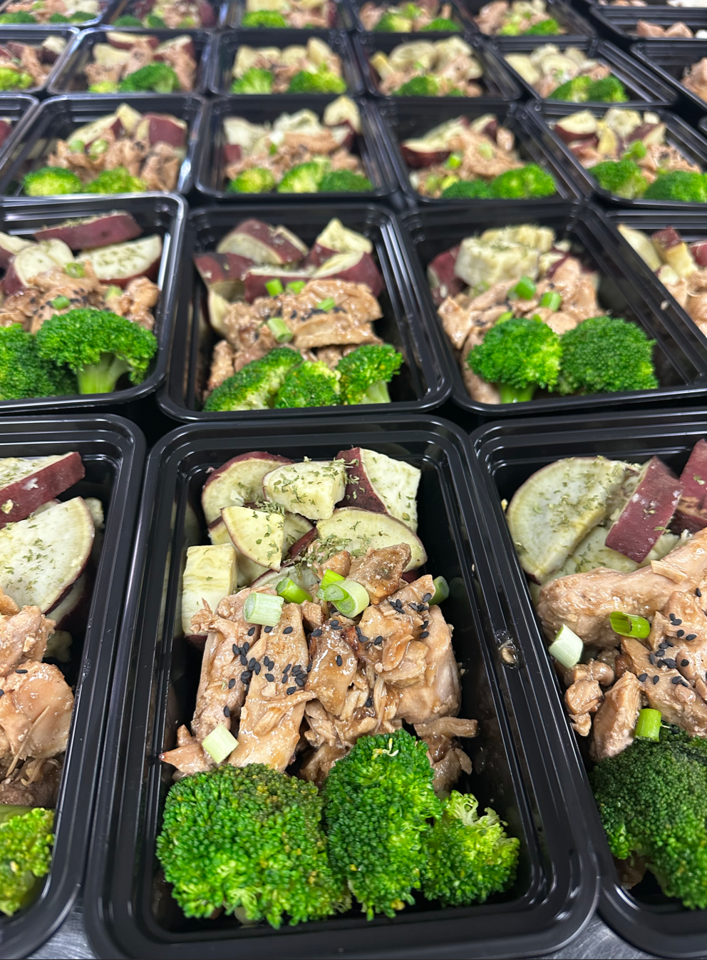 Multiple black meal prep containers filled with cooked broccoli, chopped eggplant, seasoned chicken, and diced green onions.