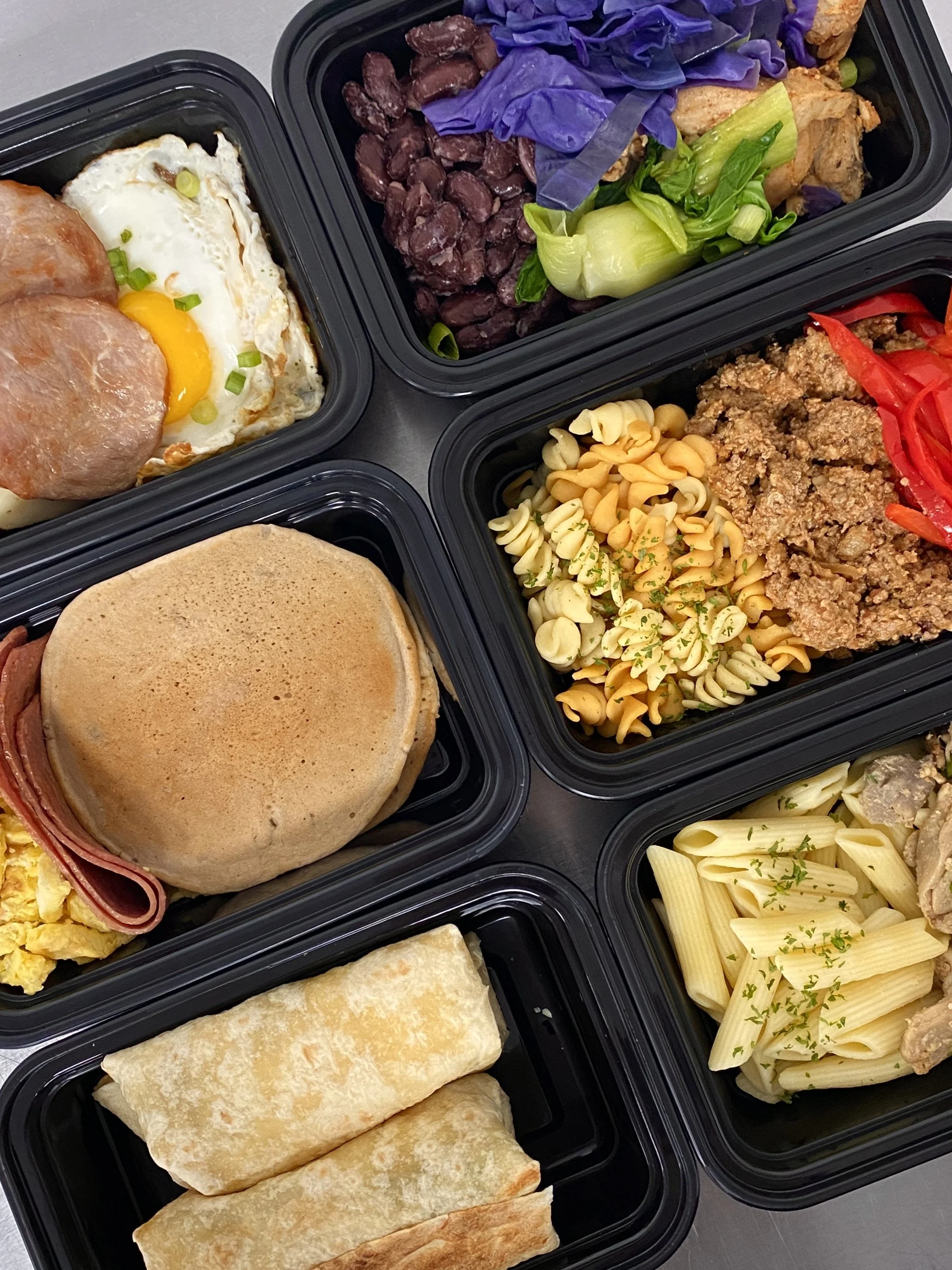 Meal prep containers with various foods including fried eggs with ham and green onions, black beans with purple cabbage and green onions, pasta salad with parsley and red bell pepper, chocolate chip cookies, a sandwich bun, a breakfast croissant wrap, and penne pasta with herbs and chicken.