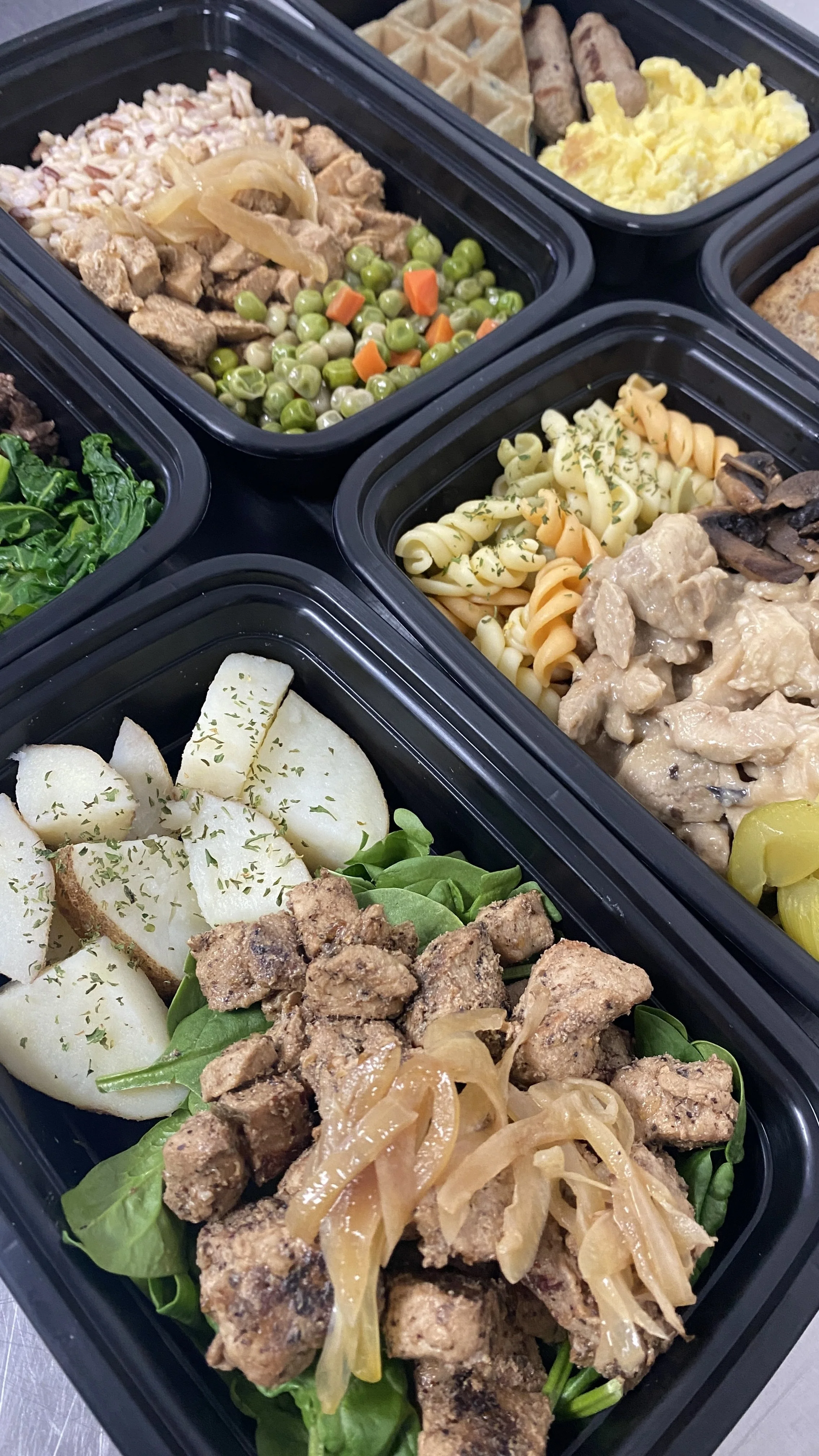 Multiple black meal prep containers filled with assorted foods including broccoli, green peas with carrots, pasta with herbs and chicken, sliced pears with herbs, and cooked chicken with onions and pickles.
