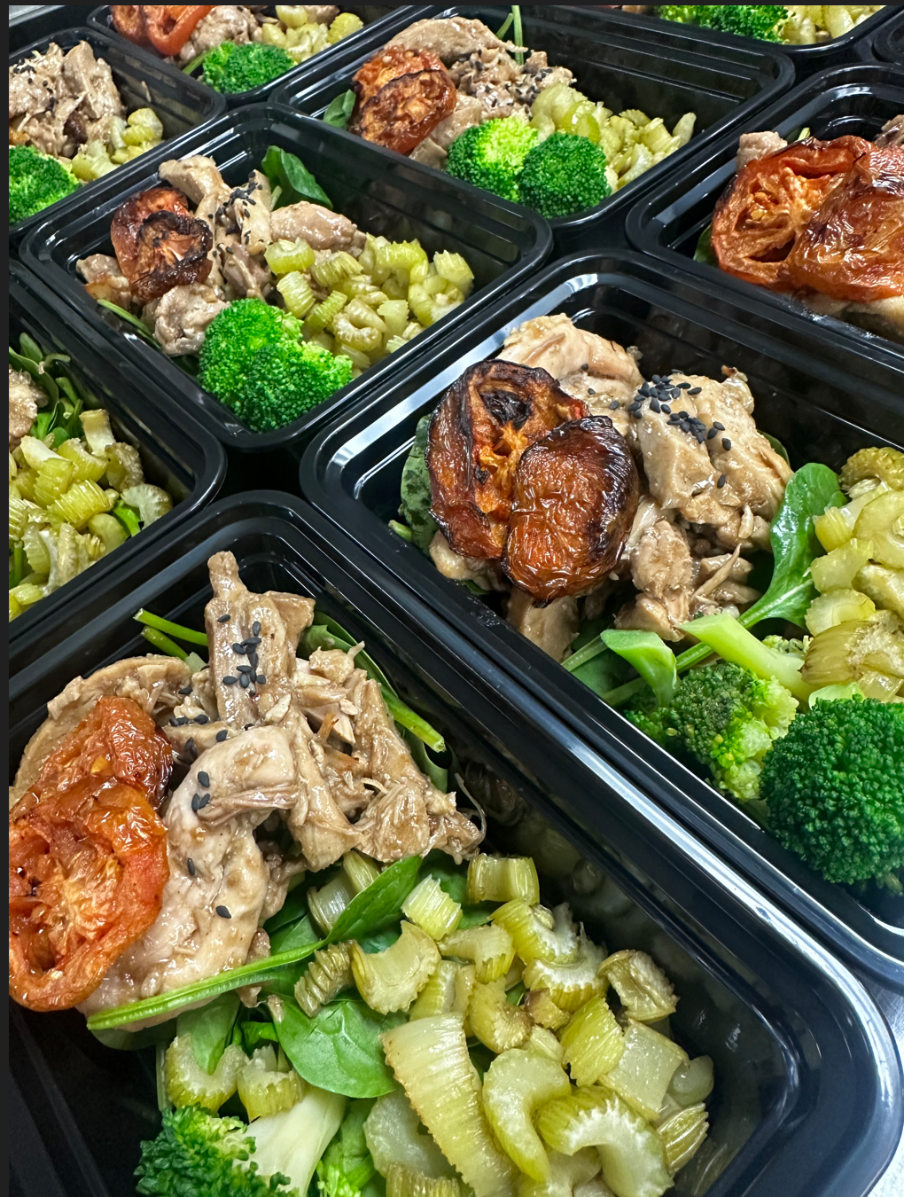 Meal prep containers filled with cooked chicken, roasted tomatoes, broccoli, celery, and greens, garnished with black sesame seeds.