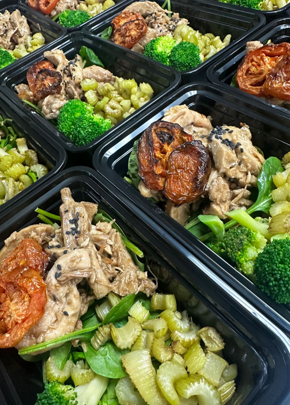 Meal prep containers with cooked chicken, roasted tomatoes, broccoli, celery, and greens.