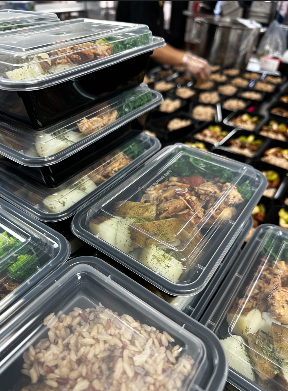 Stacked black meal prep containers filled with cooked food, with more containers in the background on a kitchen countertop.