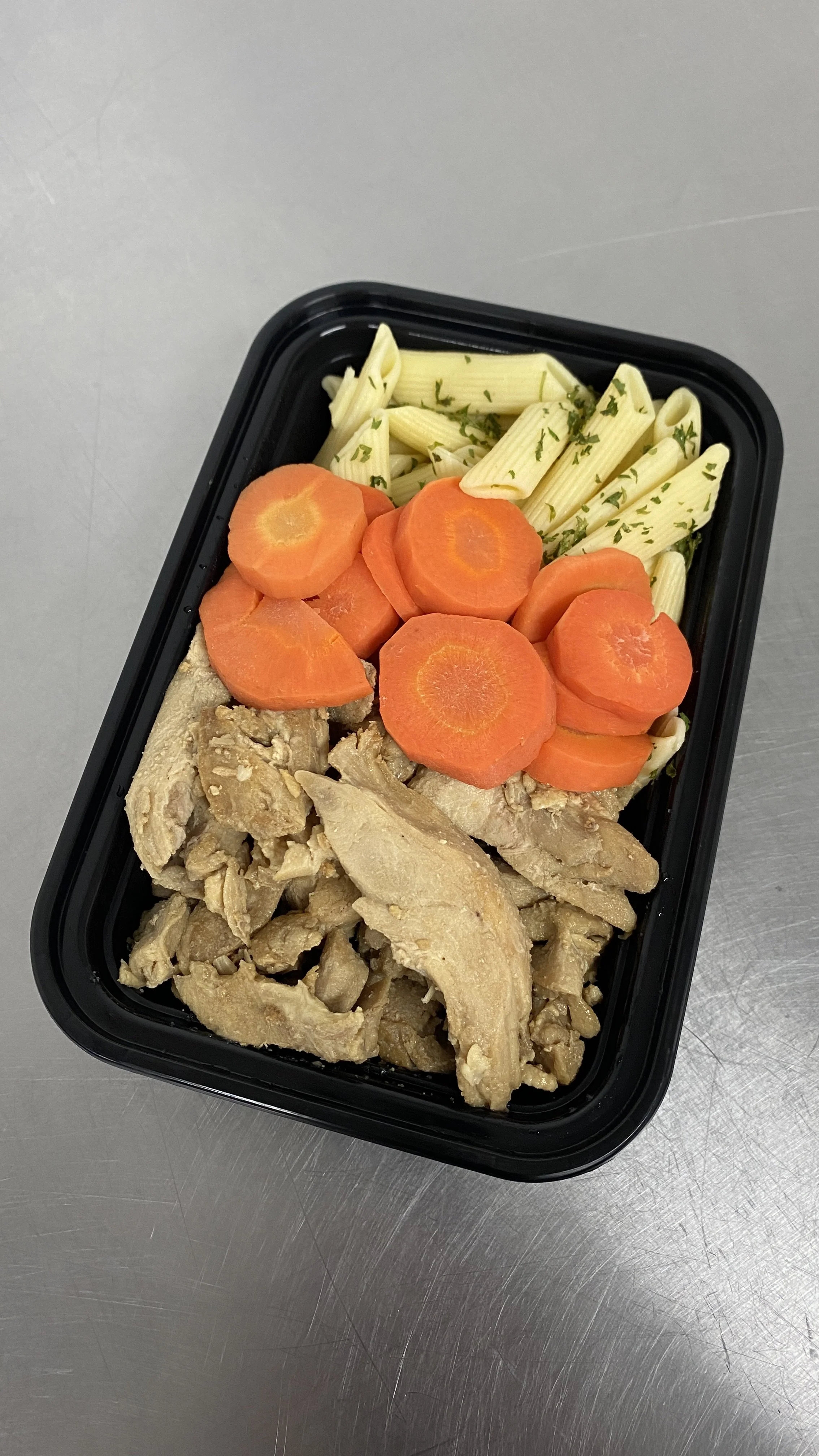 Pre-packaged meal with cooked sliced carrots, penne pasta with herbs, and cooked chicken pieces in a black plastic container.