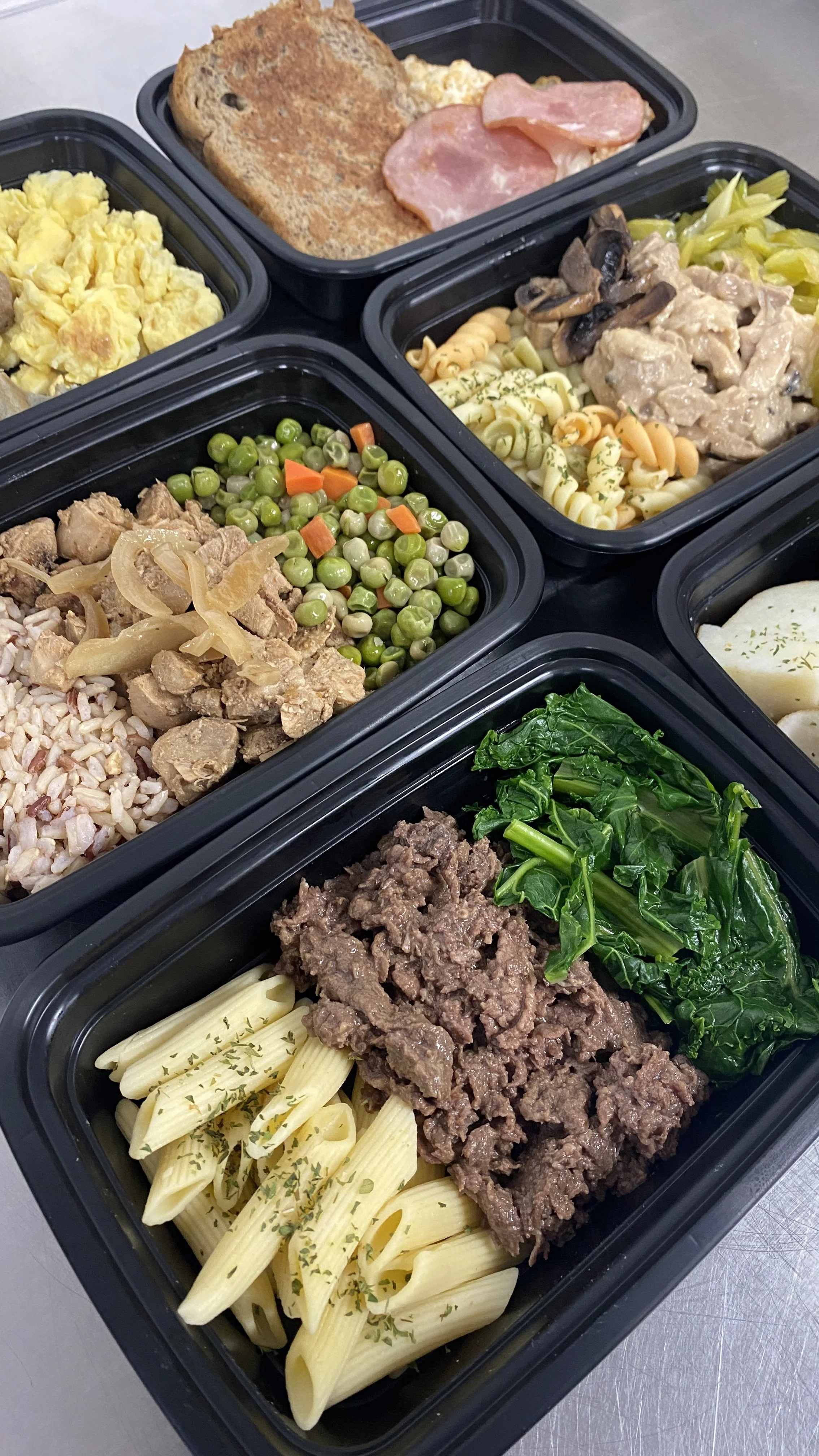 Black meal prep containers filled with pasta, ground beef, sautéed greens, peas with carrots, a creamy mushroom chicken dish, scrambled eggs, toasted bread with ham, and potato salad.