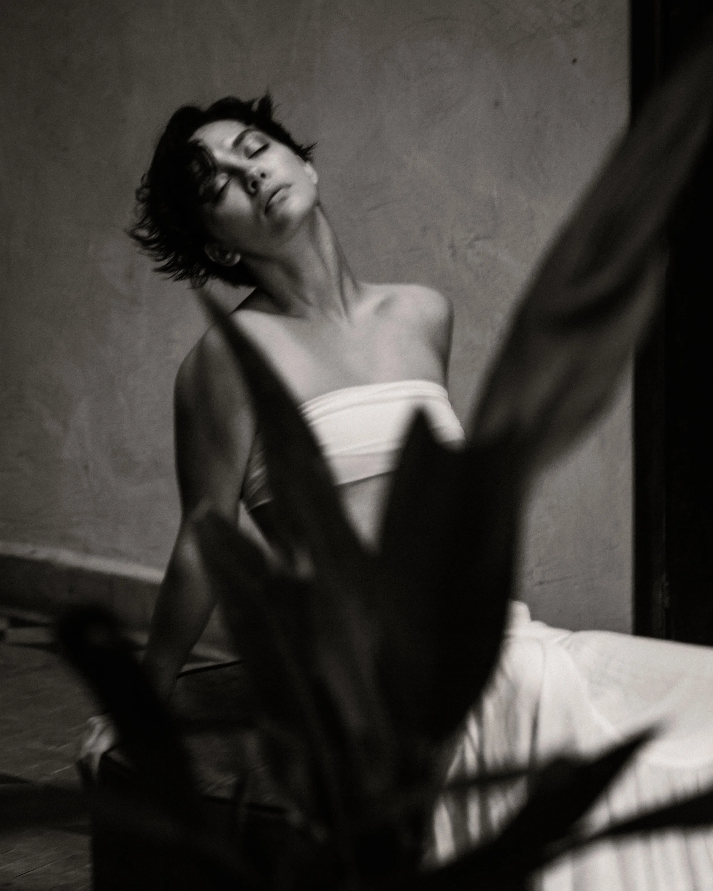 A black and white photo of a woman with short, tousled hair, wearing a strapless top, leaning against a textured wall with her eyes closed and her head slightly tilted.