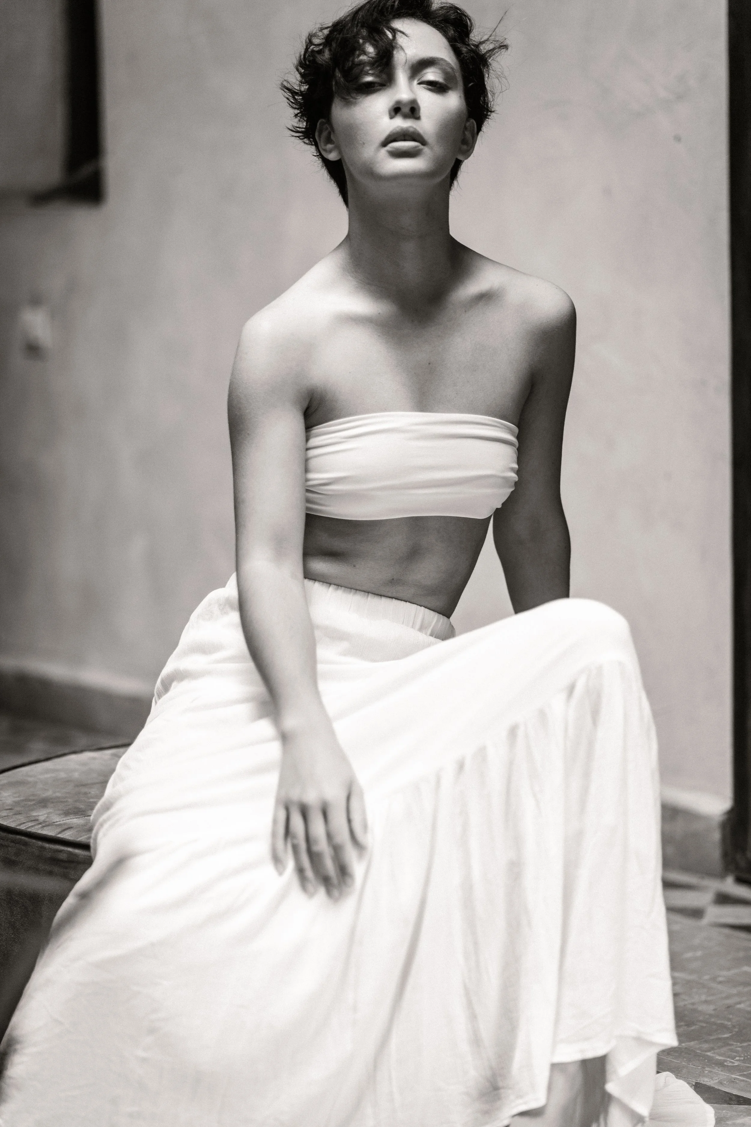 A black and white photograph of a woman with short, curly hair. She is wearing a strapless top and a long skirt, sitting outdoors on a surface with her gaze directed at the camera. Her expression is serious and her lips are slightly parted.