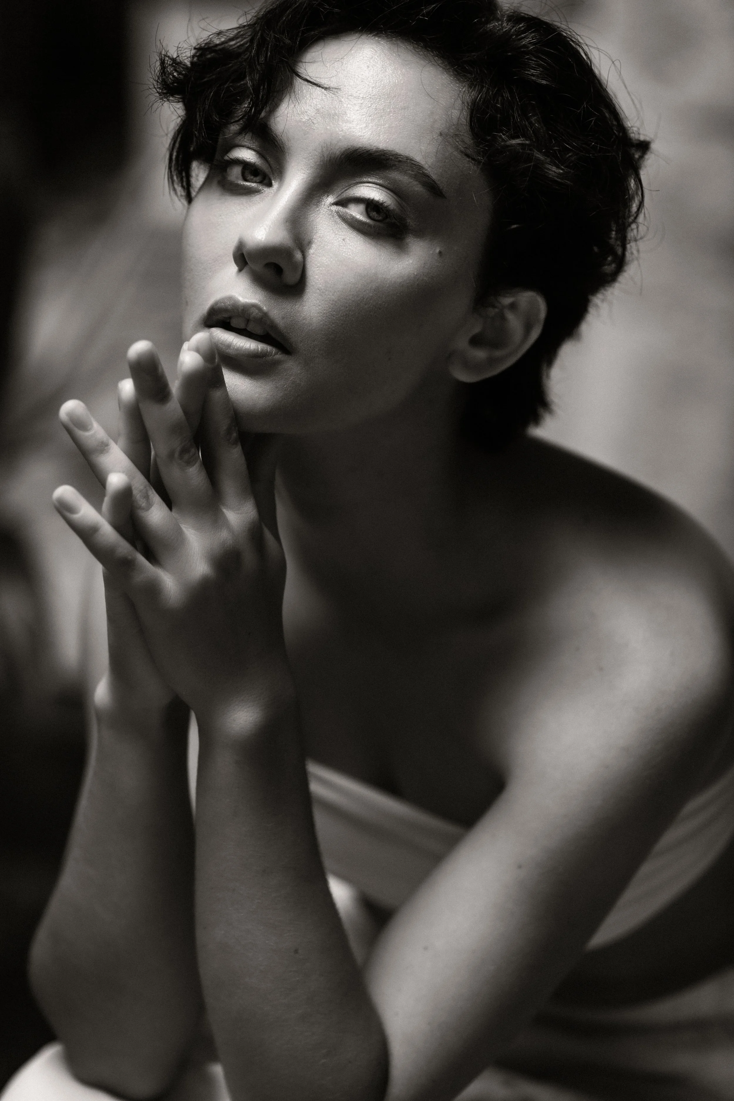 Black-and-white photograph of a woman with short, tousled hair showing her face and hands near her face, with an intense gaze and slightly parted lips.