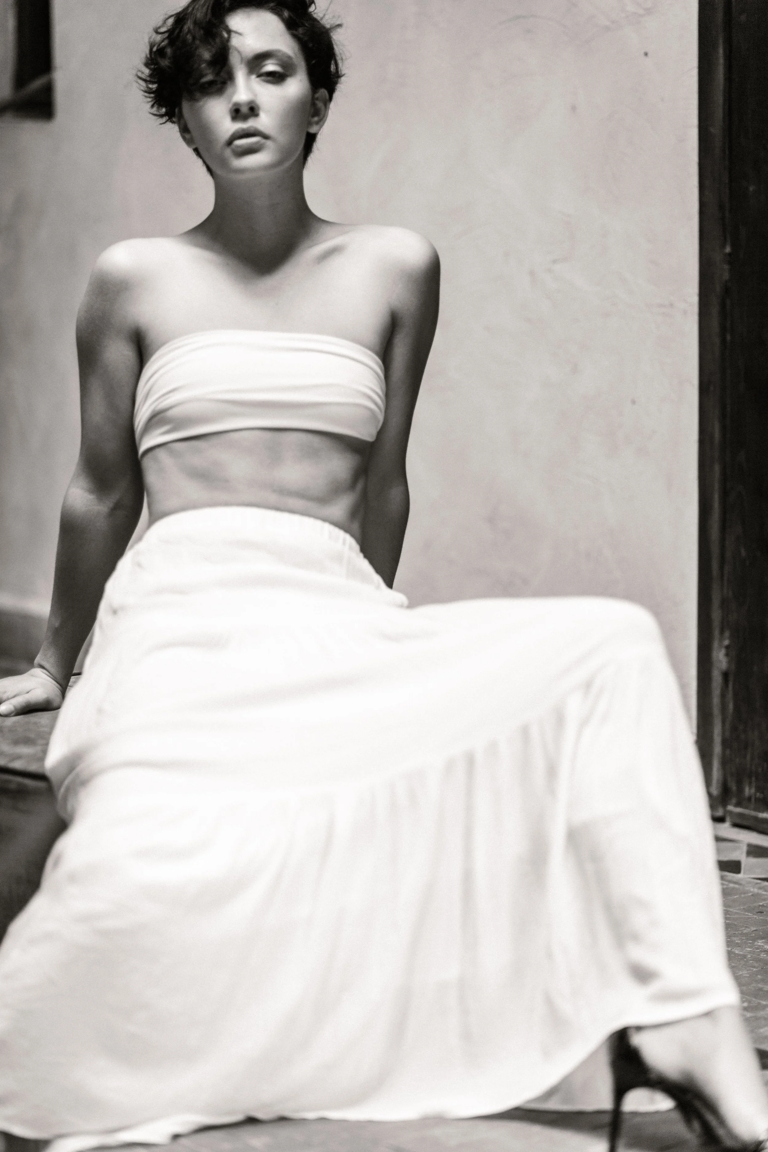 A black and white photo of a woman with short curly hair, sitting on the floor against a plain wall. She is wearing a strapless top and a long skirt, with a serious expression on her face.