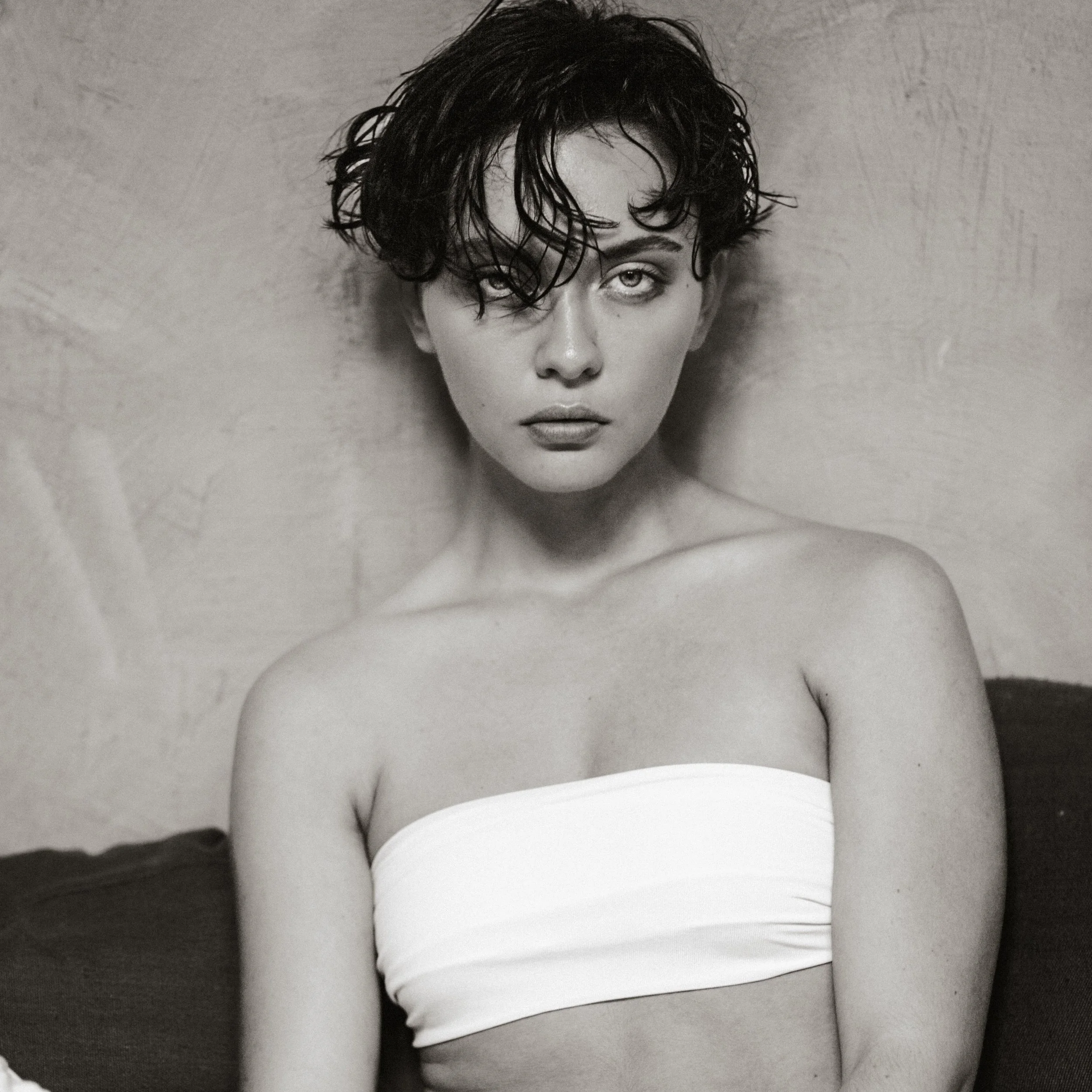 A black and white close-up photograph of a young woman with wet, curly hair, highlighting her eyes and lips, with her bare shoulders and neck visible.