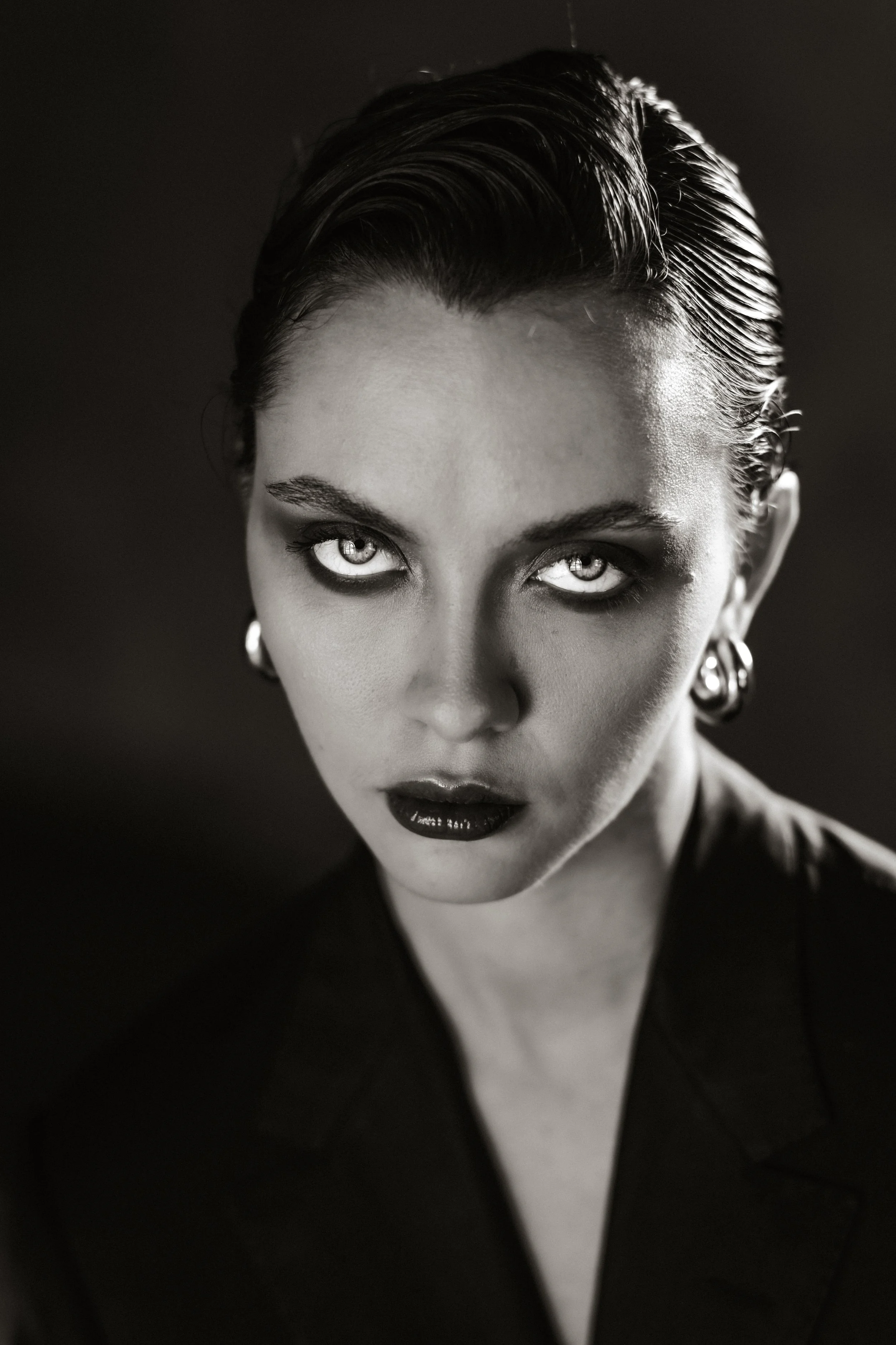 Close-up black and white portrait of a woman with slicked back hair, intense eyes, dark lipstick, and large hoop earrings, wearing a dark V-neck top.