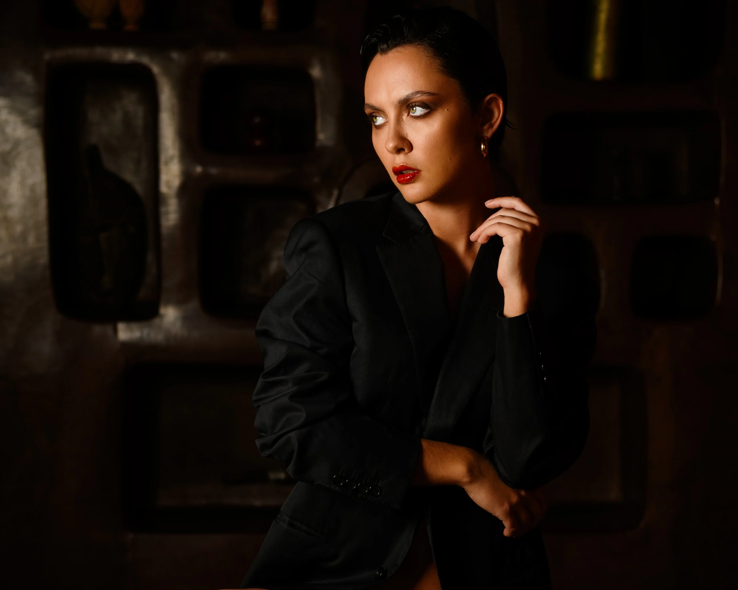 A woman with short dark hair and green eyes is wearing red lipstick and gold earrings, dressed in black, posing with her hand near her face against a dark, blurry background.