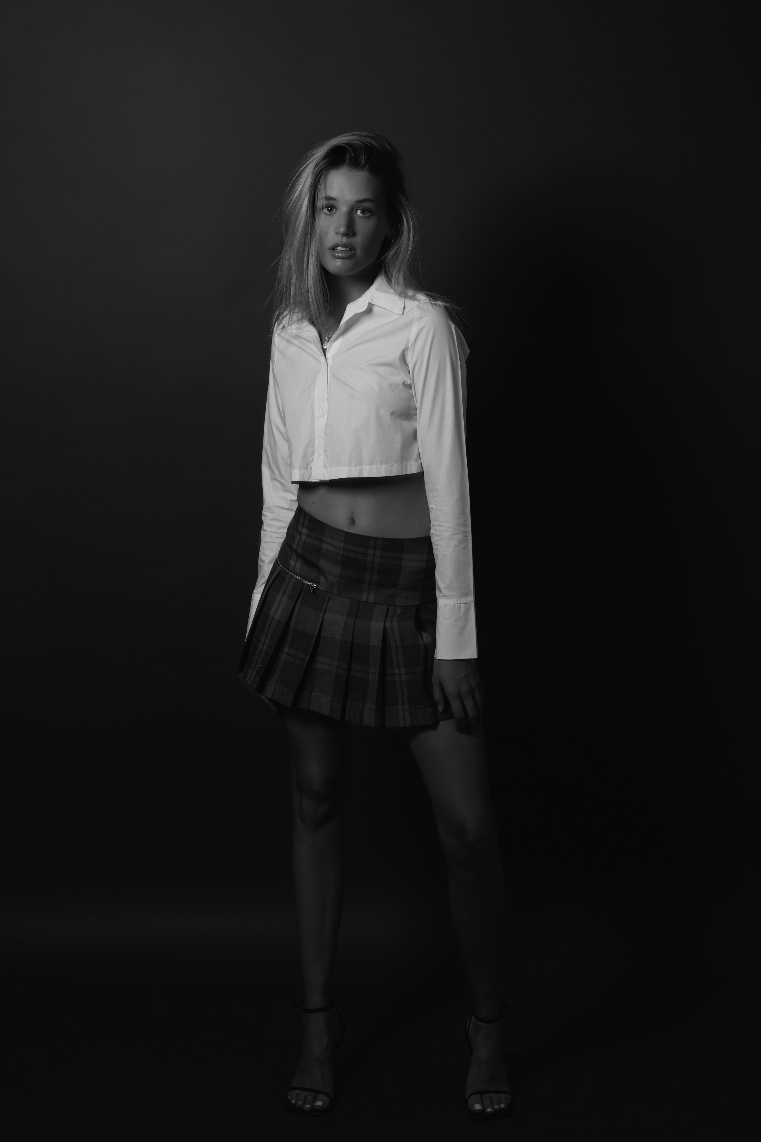 Black and white photo of a woman wearing a white cropped shirt and plaid skirt, standing against a dark background.