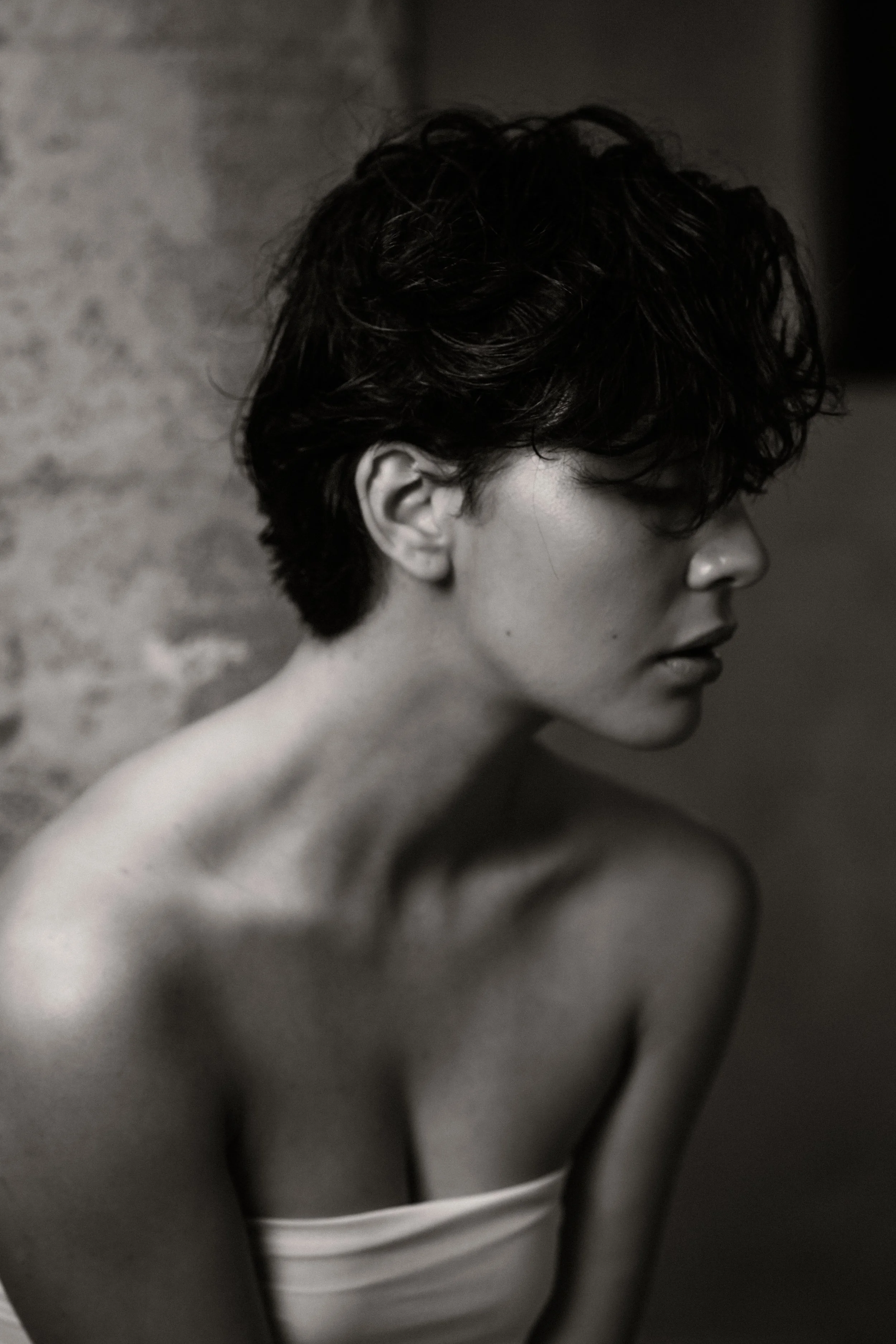 A black-and-white photograph of a woman with short, wavy hair, showing her profile from the shoulders up, with a bare shoulder and part of a white strap visible on her chest.