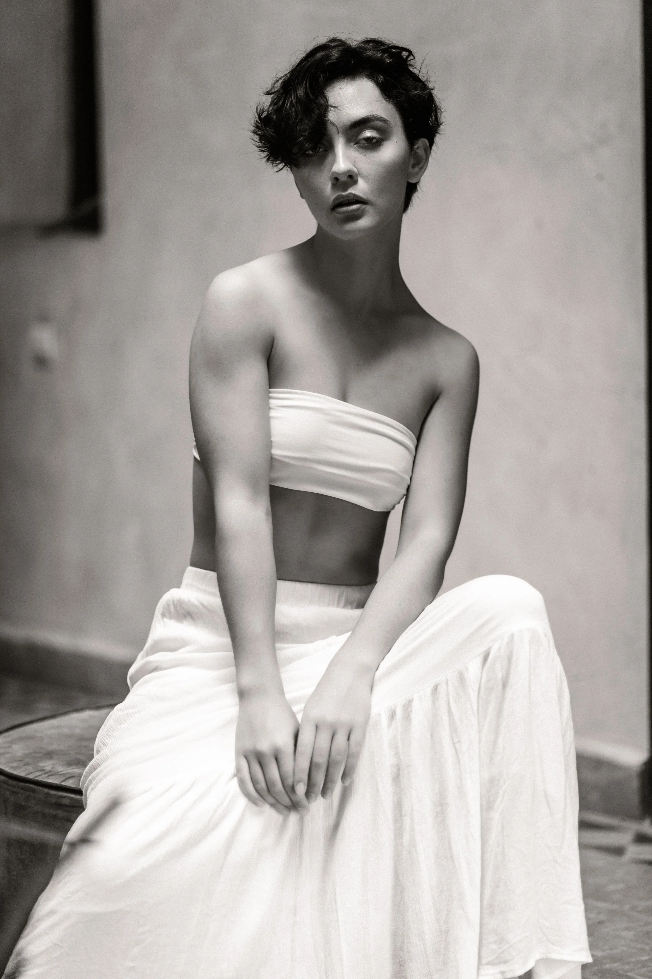 A woman with short wavy hair, sitting on a chair, wearing a strapless top and a long skirt, looking at the camera with a serious expression, in a black and white photo.