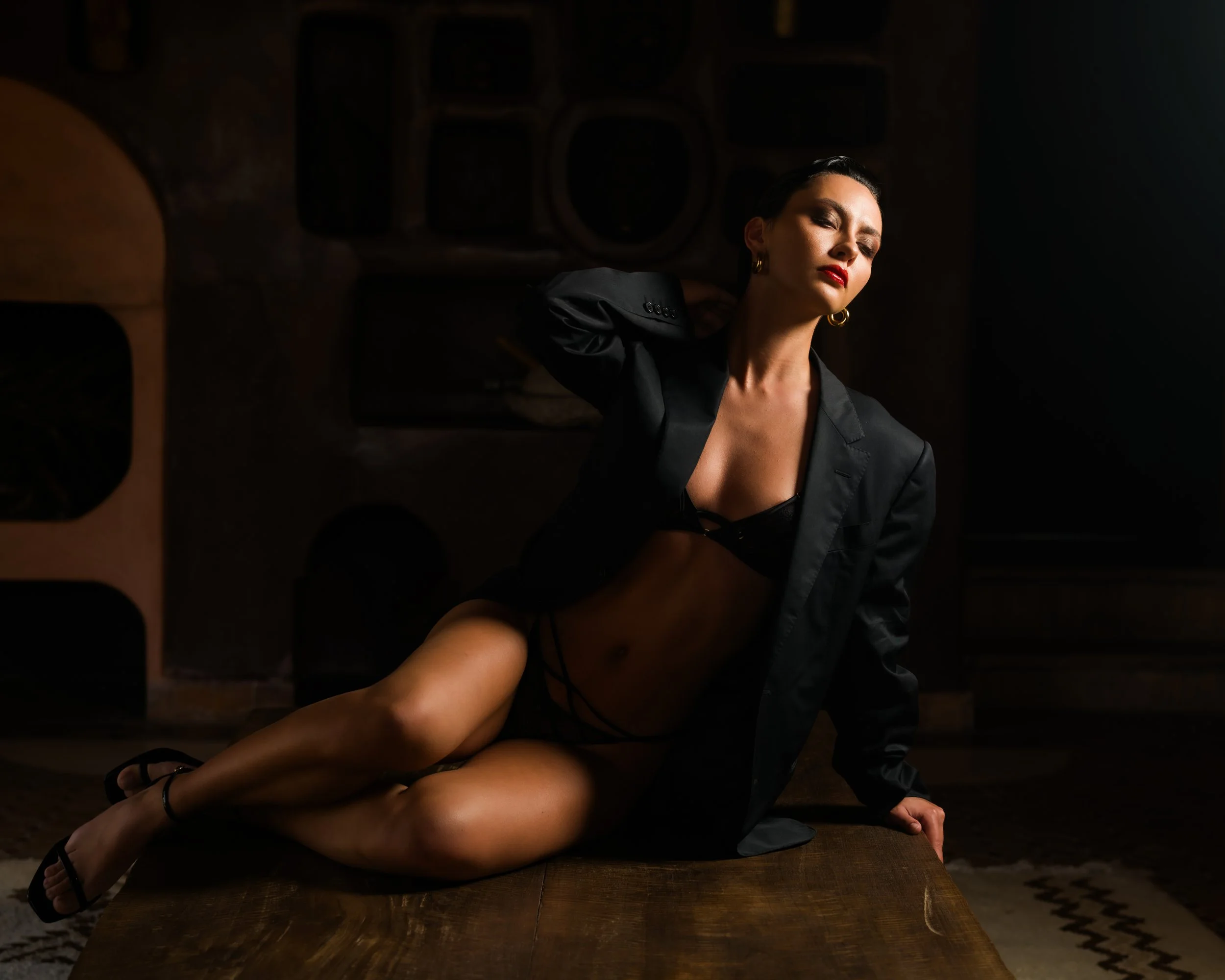 A woman wearing a black blazer and lingerie posing on a wooden table in a dimly lit room with dark walls and abstract artwork in the background.