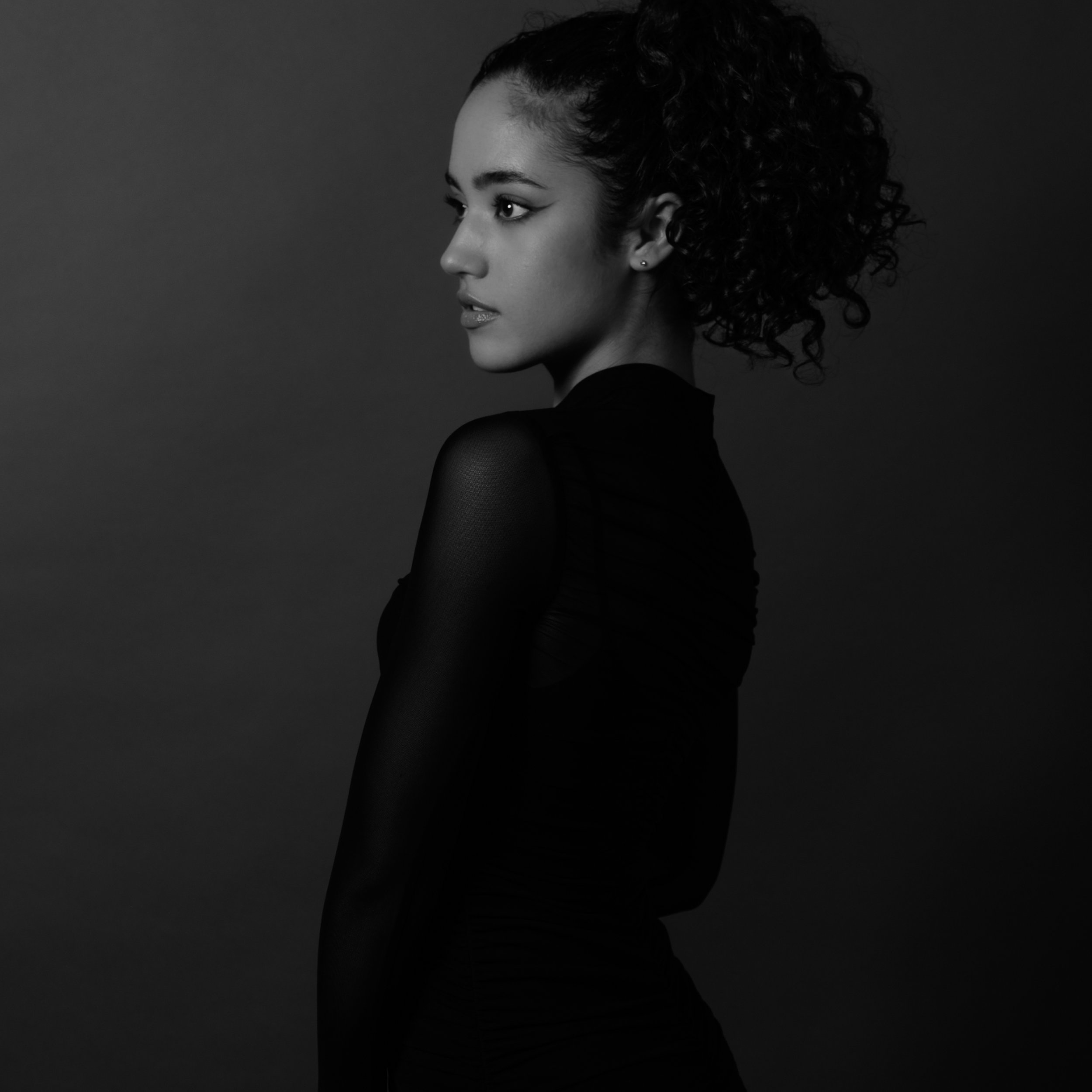 Black and white photo of a woman in profile with curly hair, wearing a dark outfit against a plain background.