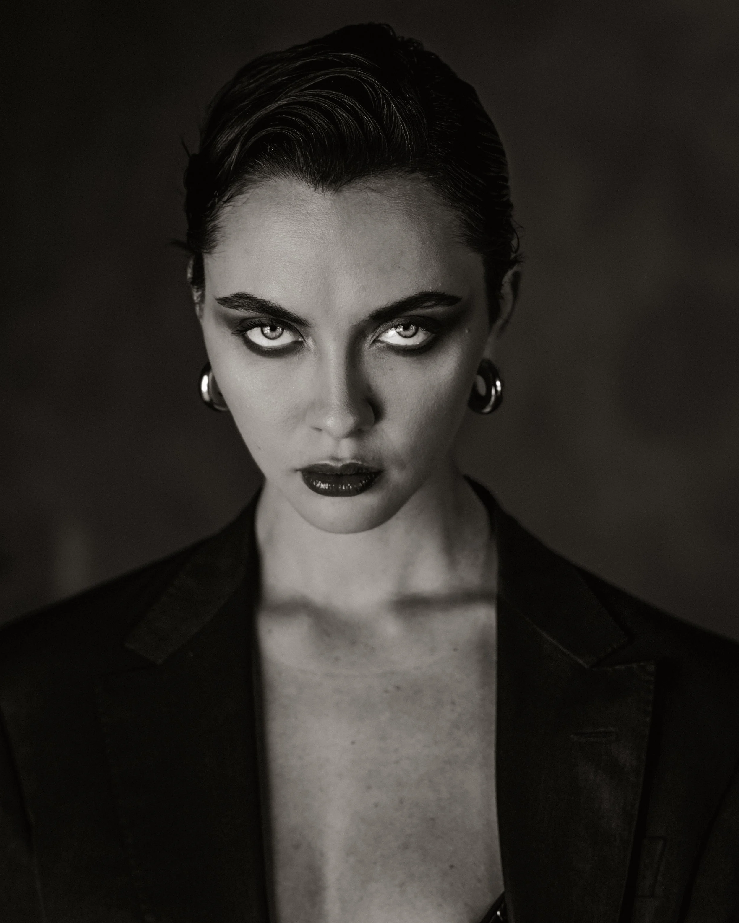 Close-up black and white portrait of a woman with short, slicked-back hair, intense gaze, dark makeup, and hoop earrings.