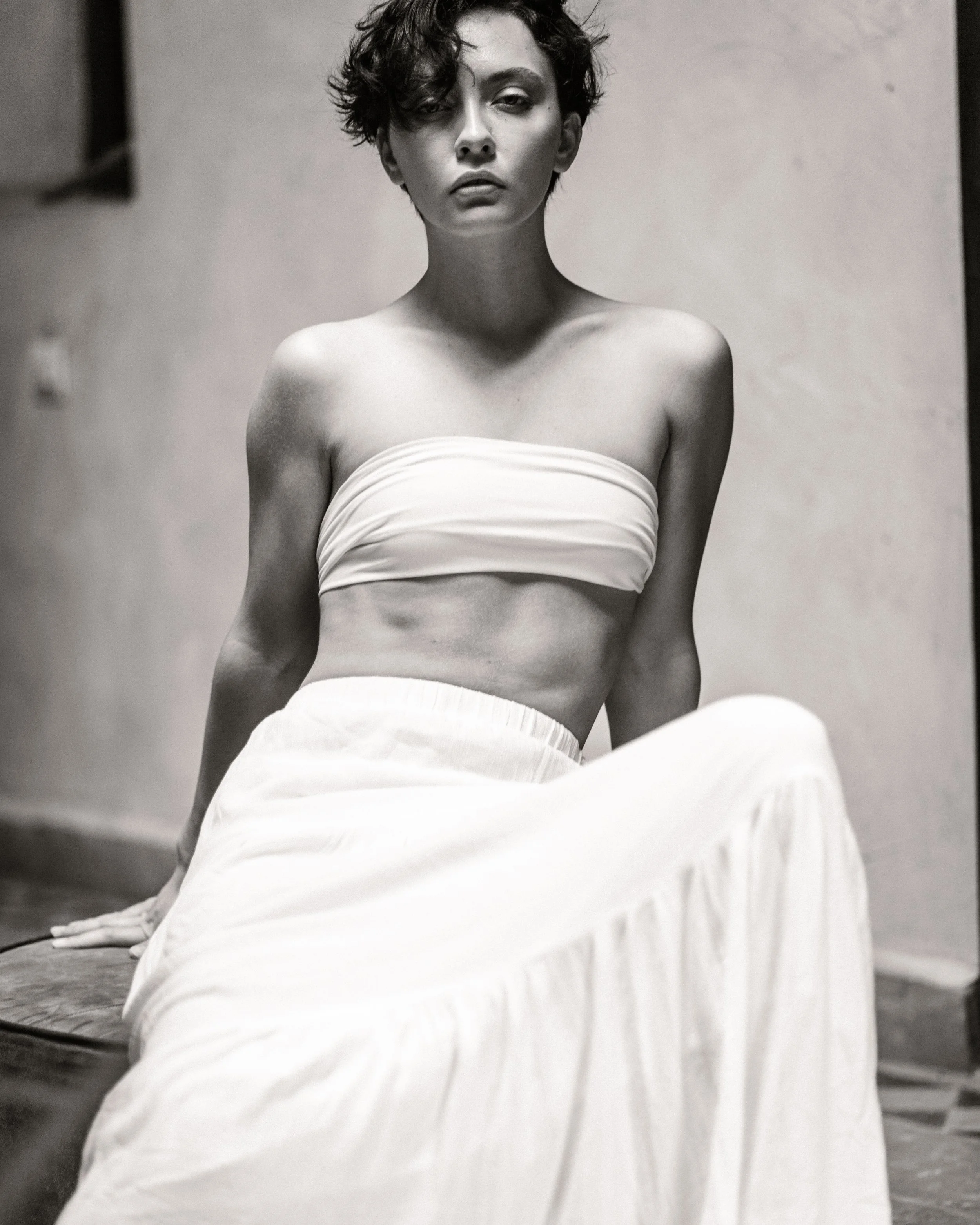 Black and white photograph of a woman with his eyes half closed, wearing a strapless top and light-colored pants, sitting on the floor against a plain background.
