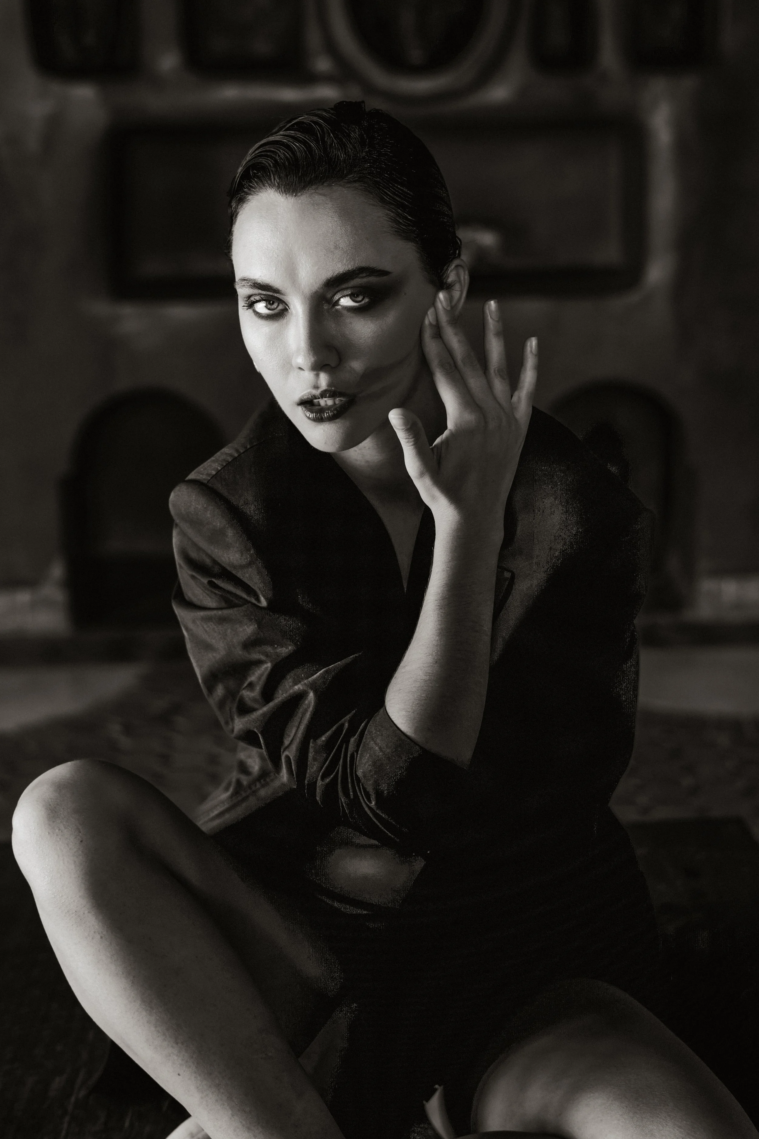 Black and white photograph of a woman with short, slicked-back hair, intense eyes, dark lipstick, and wearing a dark jacket. She is sitting with one leg crossed over the other, with her hand near her cheek, in front of a fireplace.