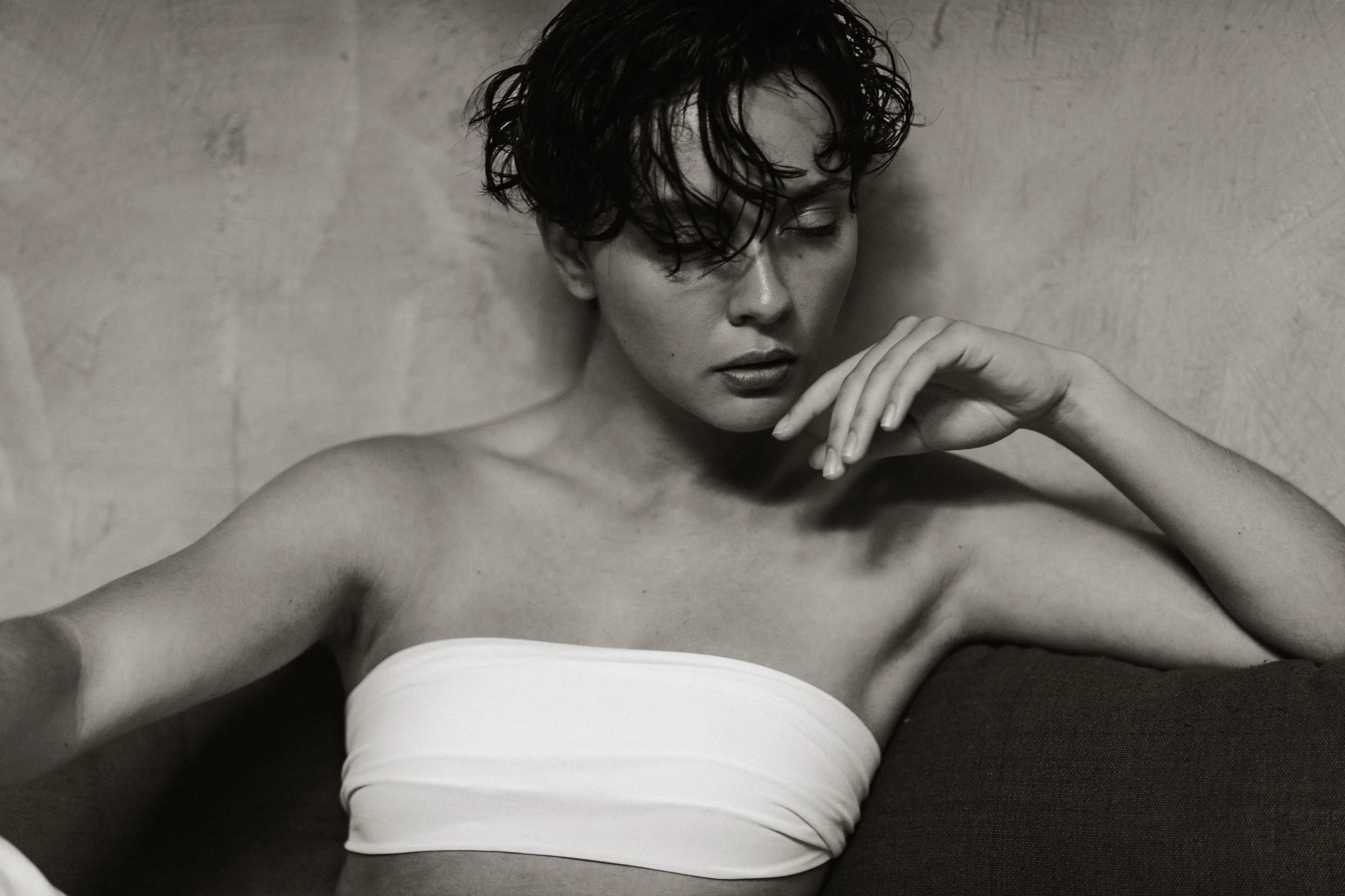 A black and white photo of a woman with wet, curly hair, closed eyes, and a contemplative expression, leaning against a textured wall, with one hand near her face.