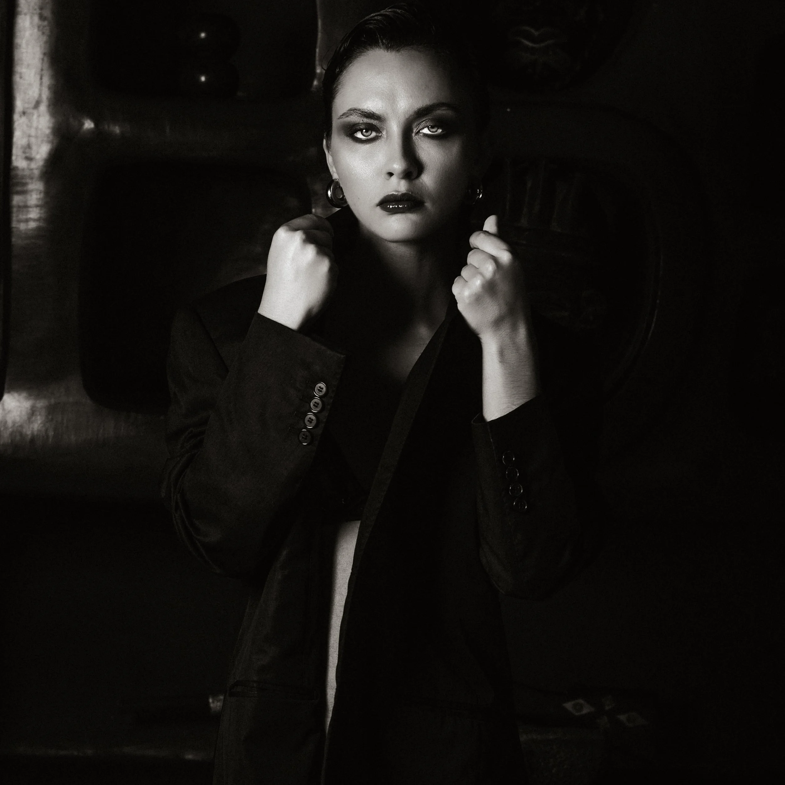 A black-and-white portrait of a woman with dark lipstick and eye makeup, holding her fists near her chin, with a dark and blurred background.