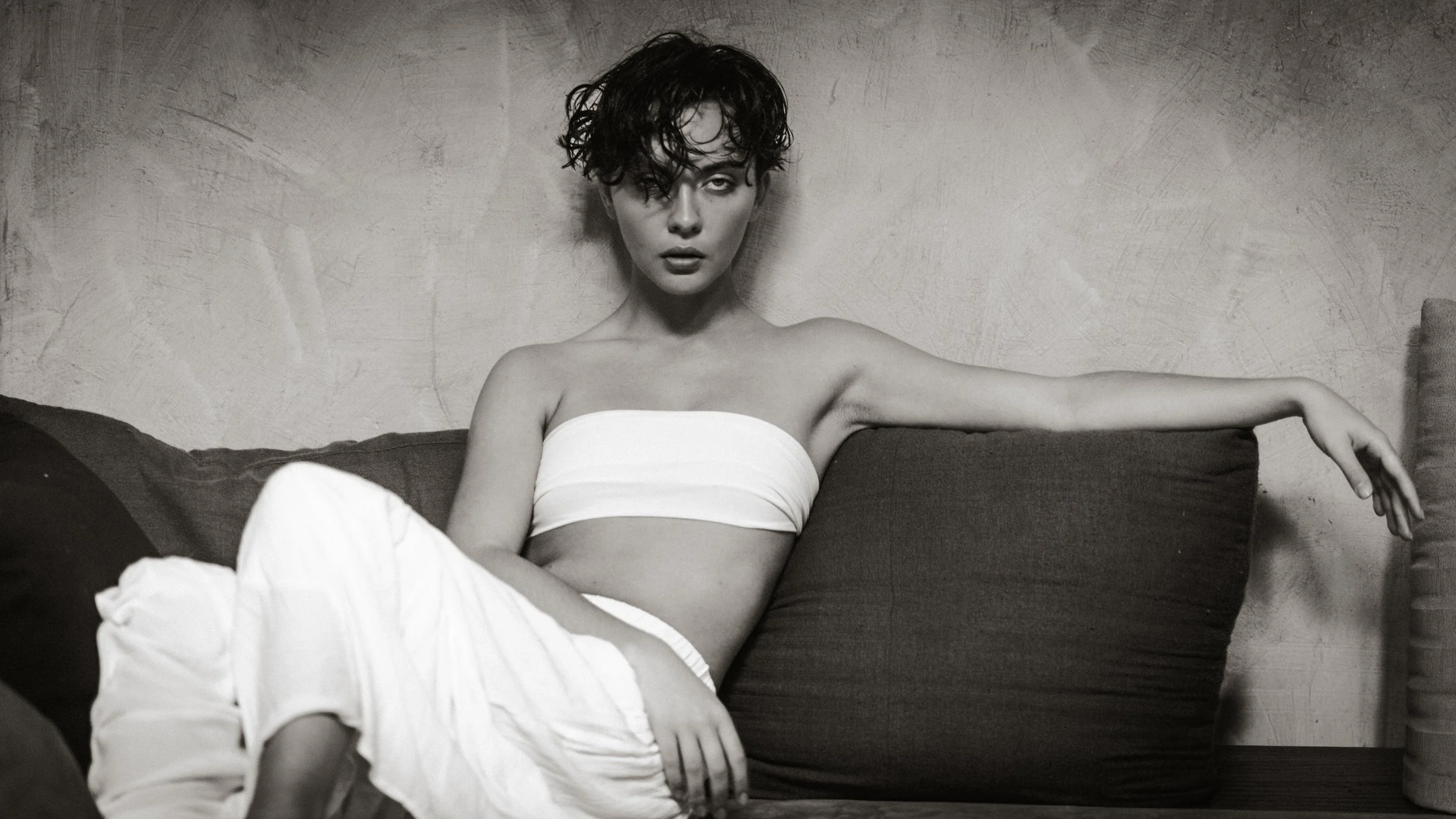 A woman with short, curly hair and minimal makeup sitting on a couch against a textured wall, wearing a strapless top and white pants, with her left arm extended along the back of the couch and her expression intense.