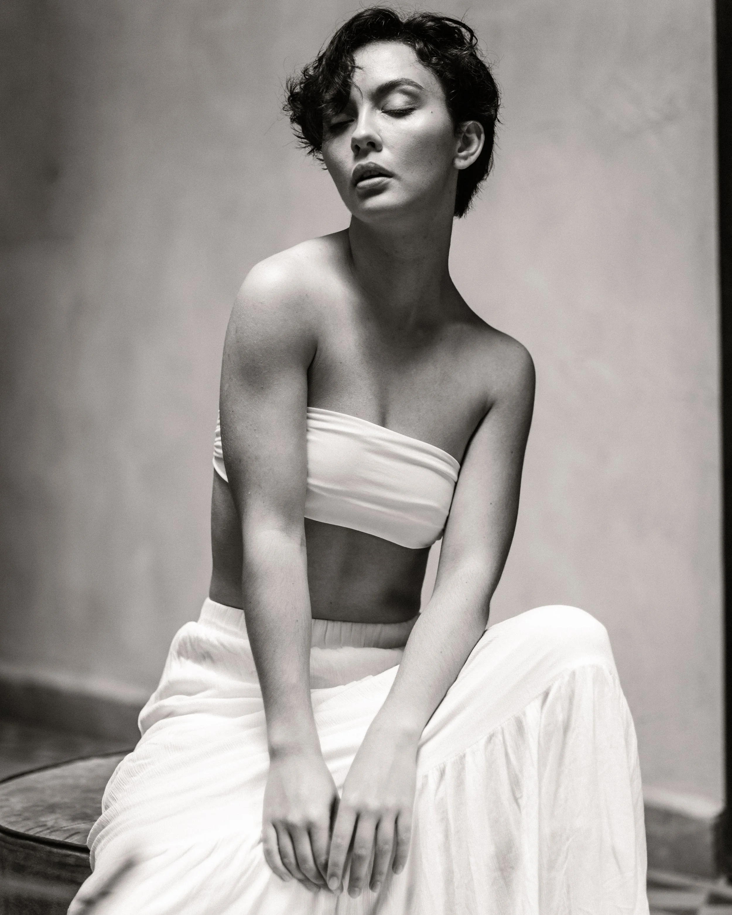 A black and white portrait of a woman with short curly hair, eyes closed, sitting with a relaxed expression, wearing a strapless top and a flowing skirt.