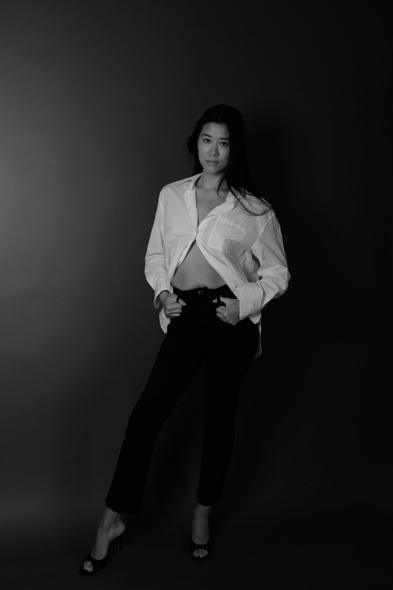 Black and white photo of a woman in a white shirt and black pants posing against a dark background.