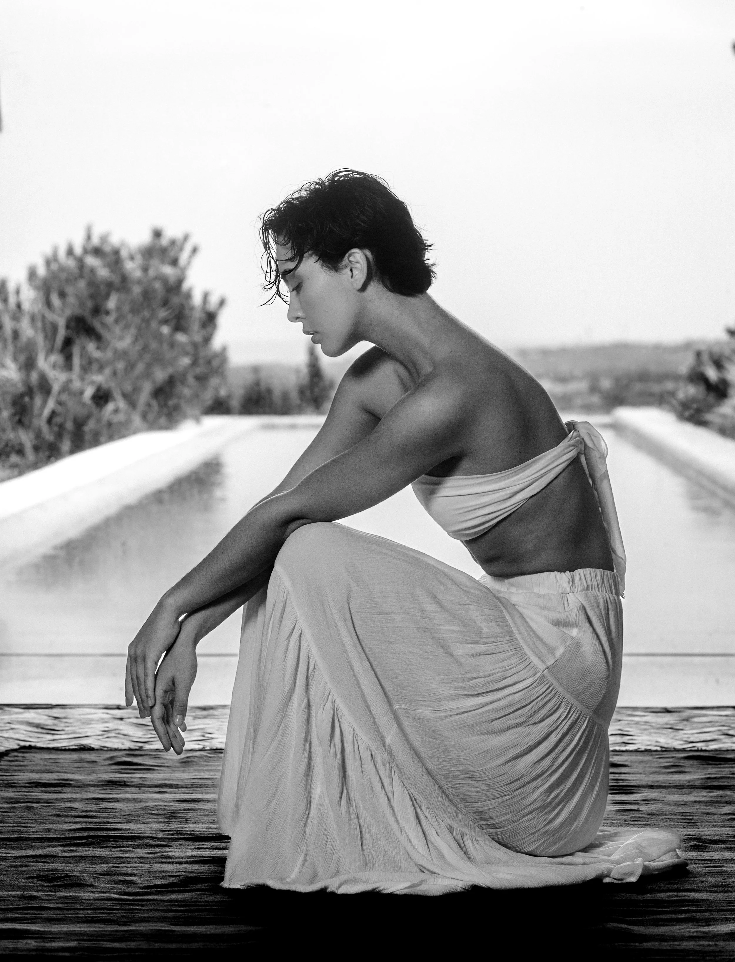 A black and white photograph of a woman sitting on a dock or pier by the water, with her head bowed and eyes closed, wearing a flowing dress and having short dark hair, with a blurred background of trees and a horizon.