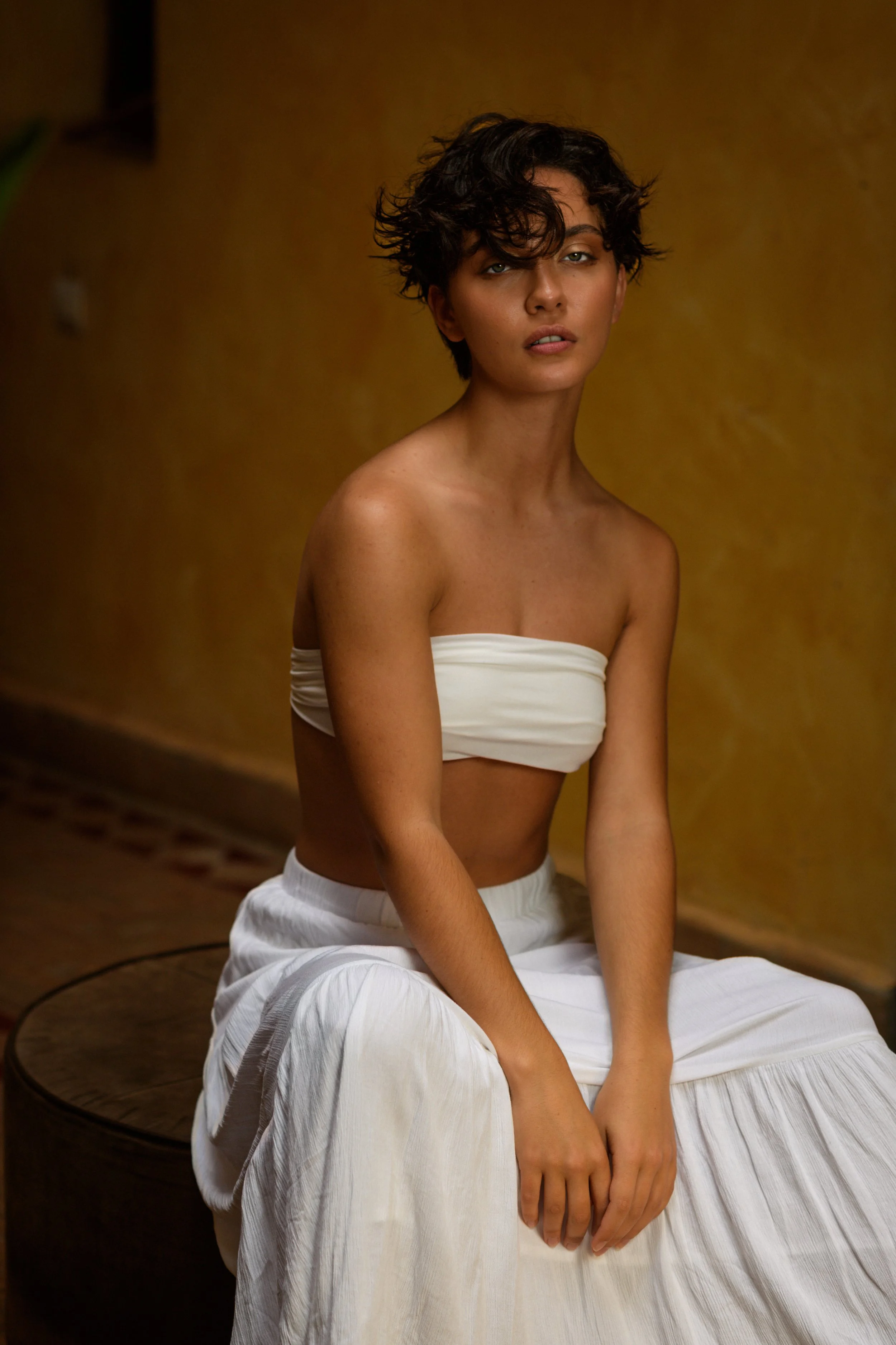 A woman with short curly hair and light skin wearing a strapless white top and white skirt, sitting on a round table with a yellowish wall in the background.