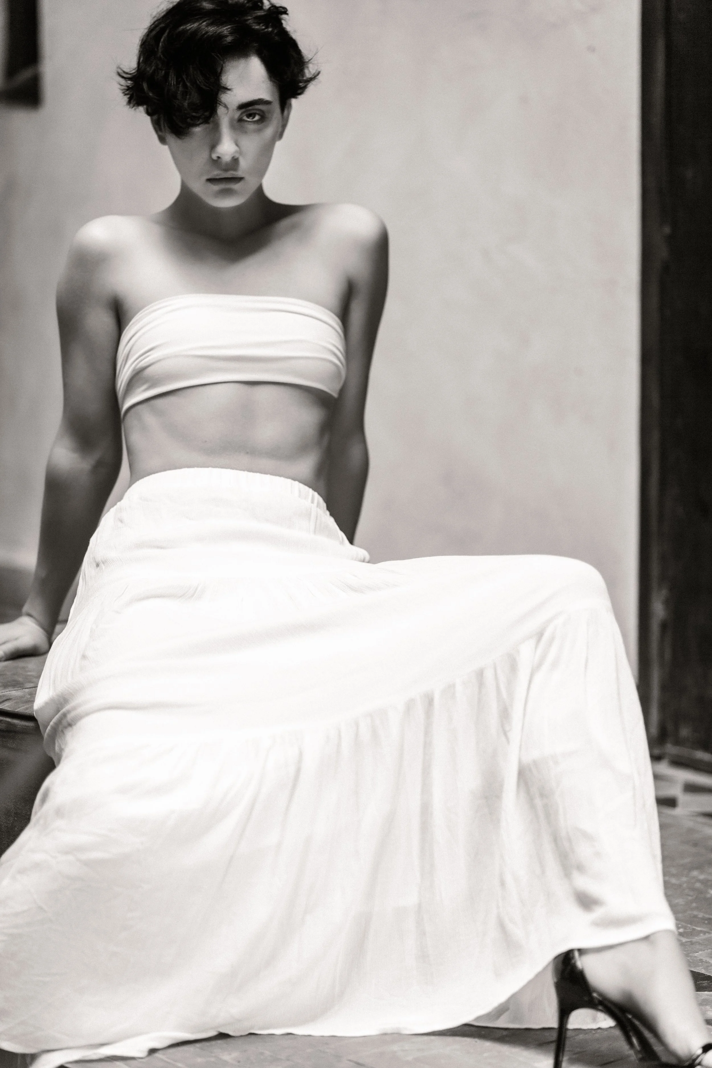 Black and white photograph of a woman sitting on the floor, wearing a strapless top and a long skirt, looking directly at the camera with a serious expression, with dark hair styled in a short, curly bob.