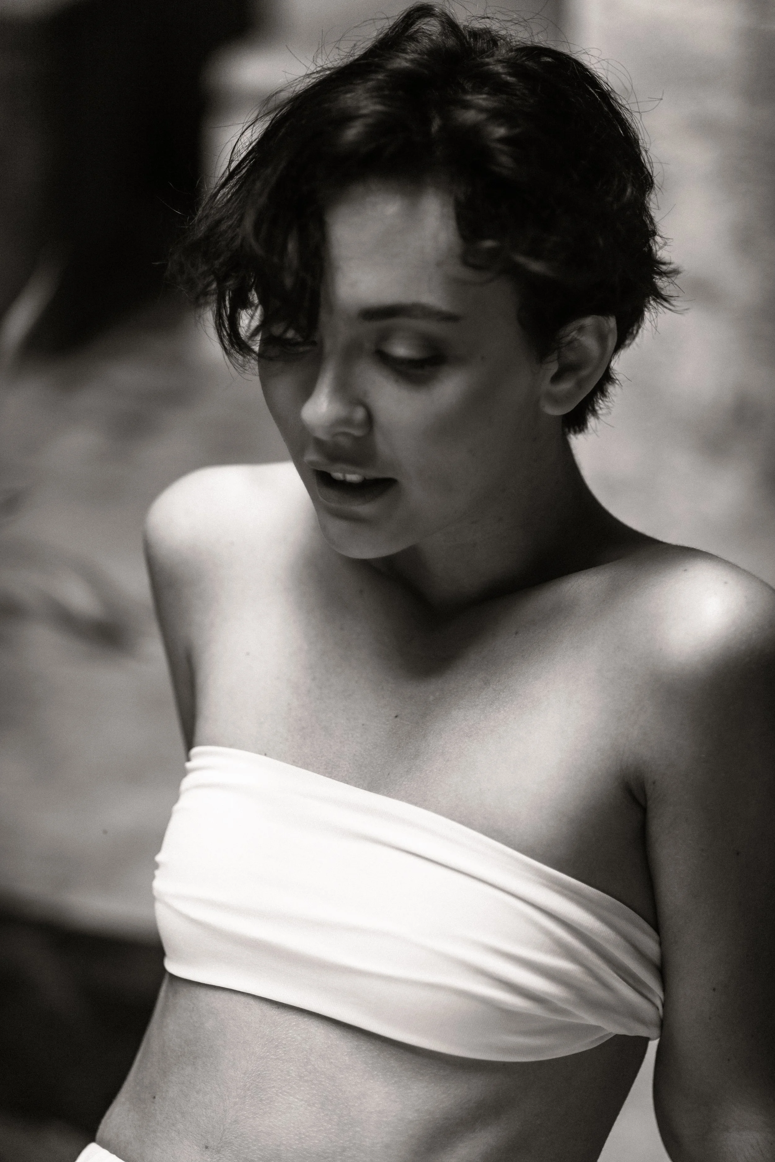Black and white photo of a woman with short, tousled hair, wearing a strapless top, with her eyes closed and mouth slightly open.