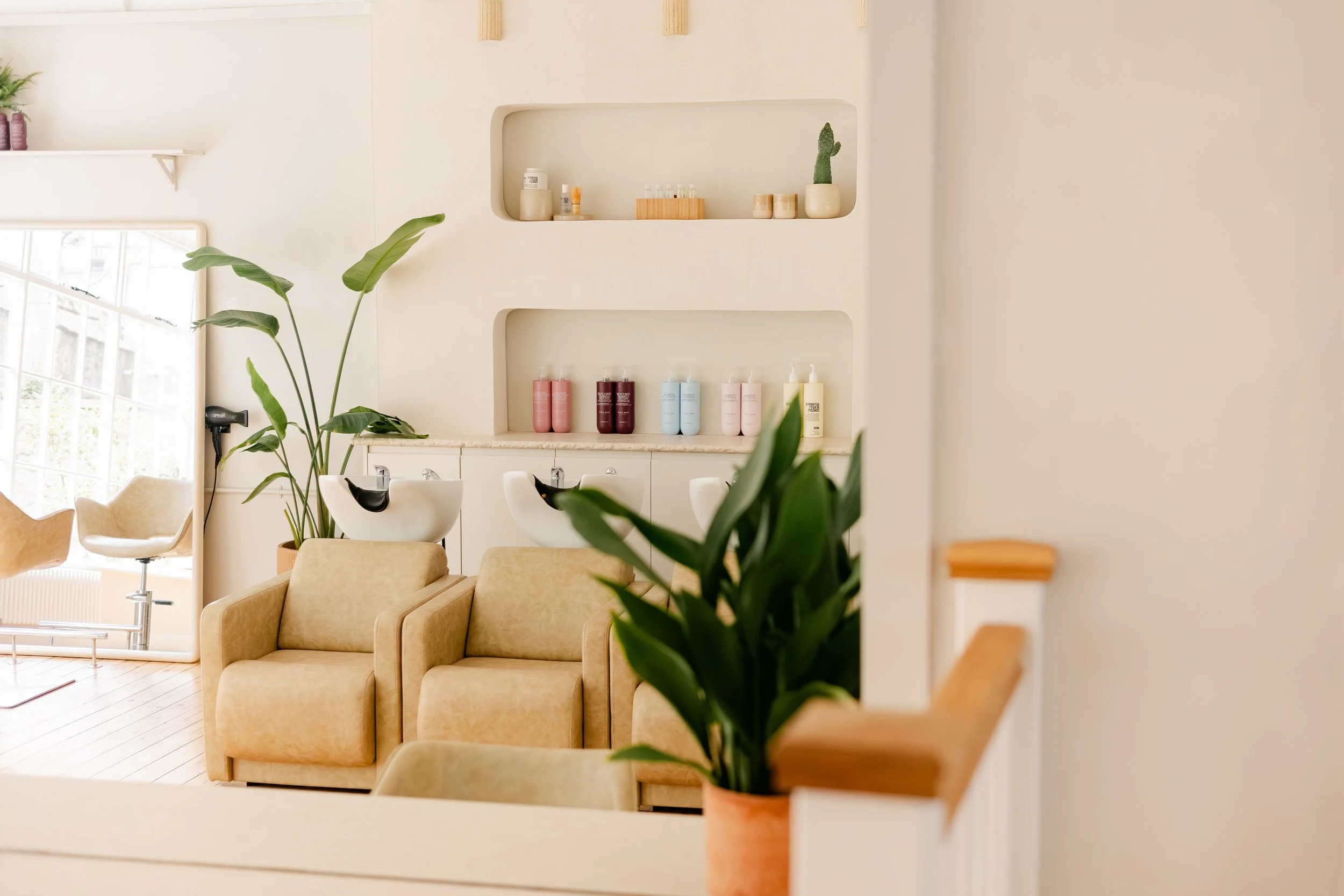 Interior of a premium, minimalistic hair salon or wellness brand with beige chairs, green plants, and shelves with skincare products and decorations.