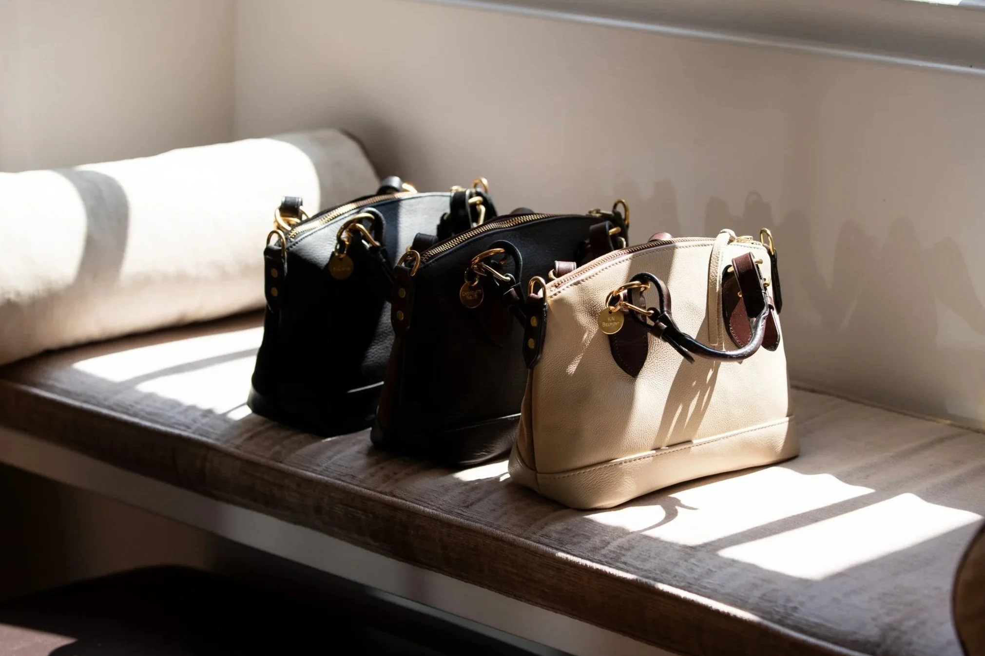 Three handbags, black, dark brown, and beige, placed on a wooden bench in sunlight. Shadows cast on the bench.
