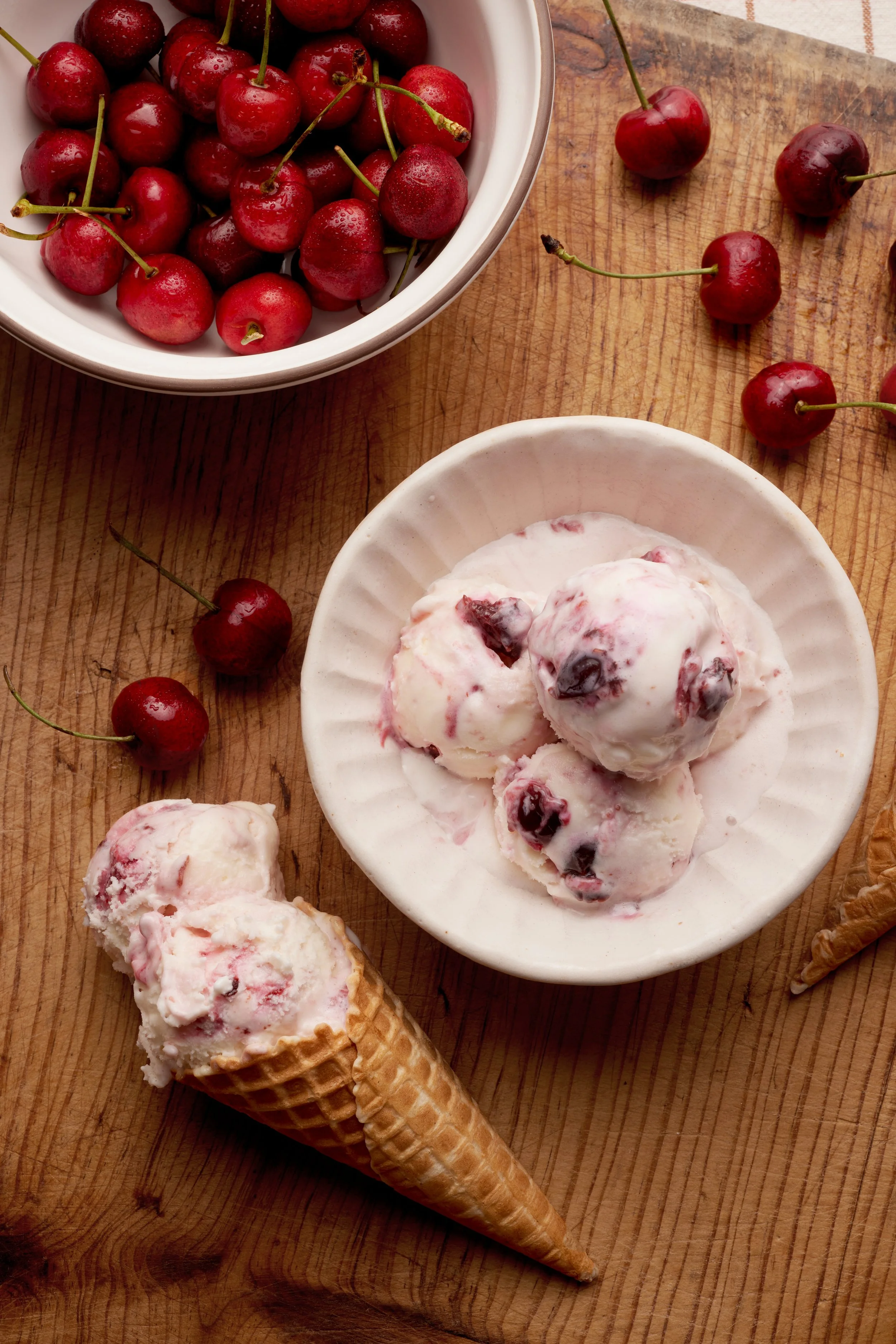 A bowl of fresh cherries, a bowl of cherry ice cream, and a cherry ice cream cone on a wooden surface.